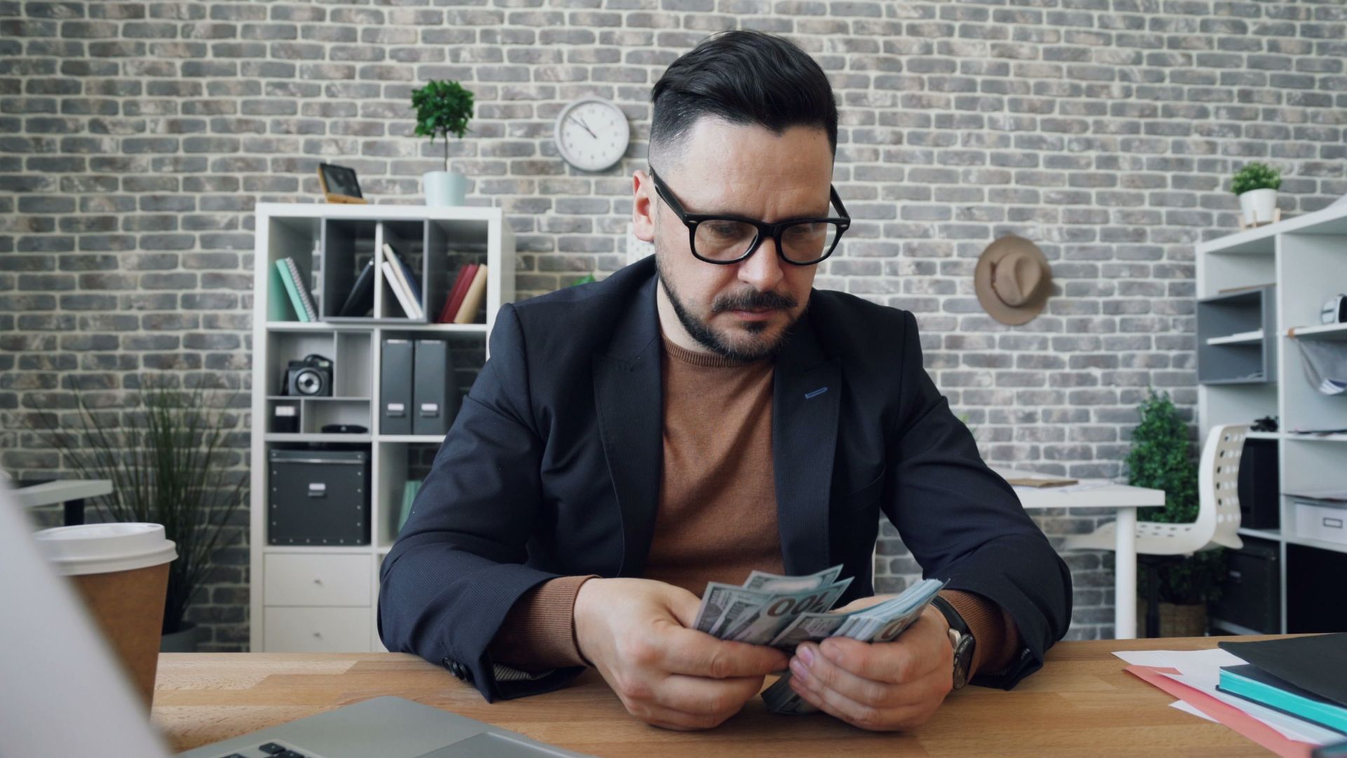 Businessman counting cash at a desk in a modern office setting with a laptop and documents.