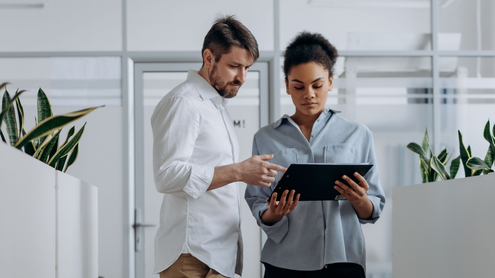 Two professionals discussing work on a tablet in a modern office hallway.