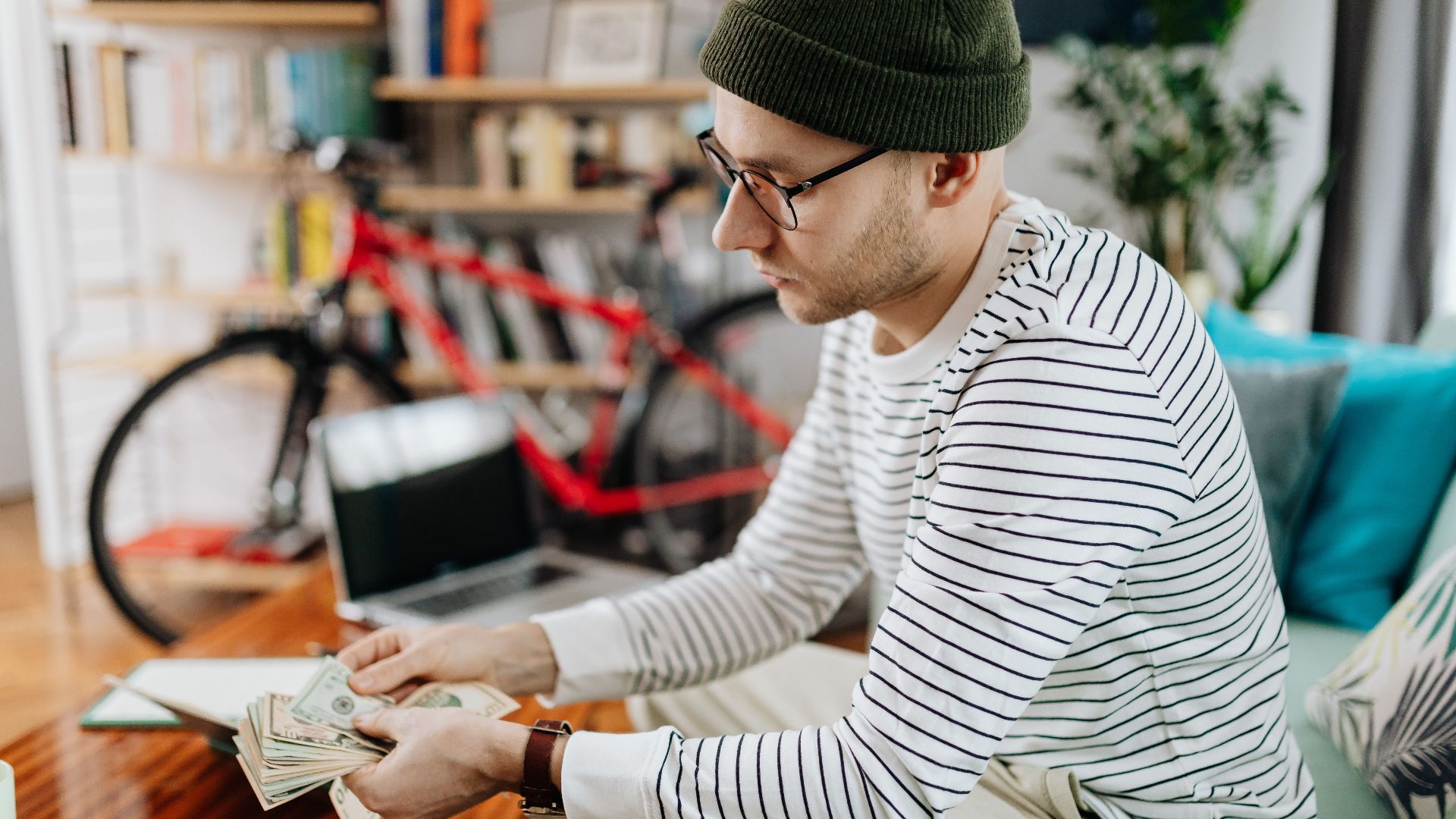 Man in striped shirt counting cash indoors, with a bicycle in the background.