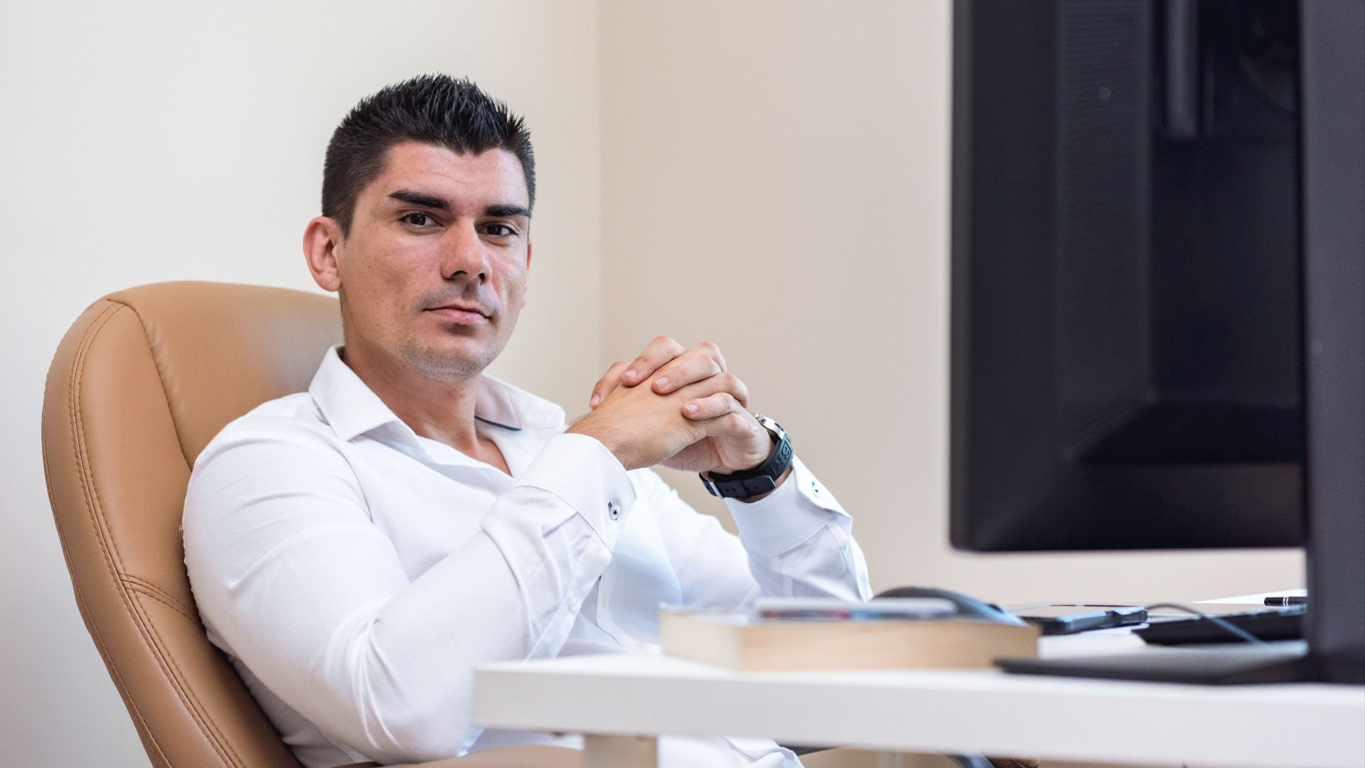 Businessman in white shirt sitting confidently at his desk with hands clasped, next to a computer monitor.