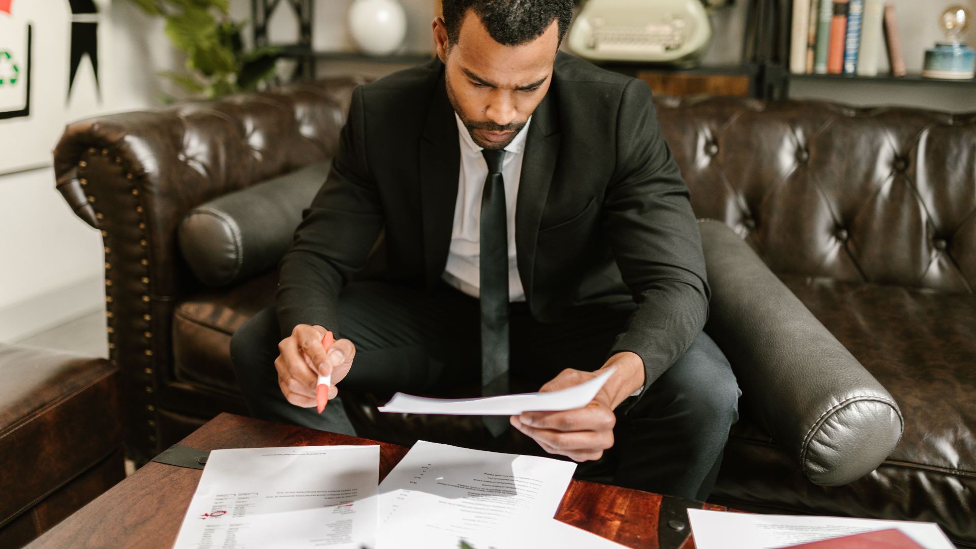 A businessman in a suit attentively reviews paperwork on a brown leather sofa.
