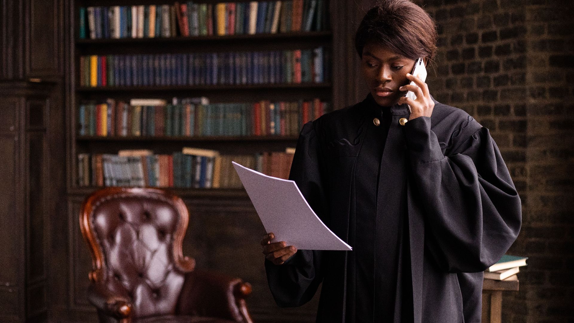 African American judge in traditional robes holds a document and talks on the phone in a law library.