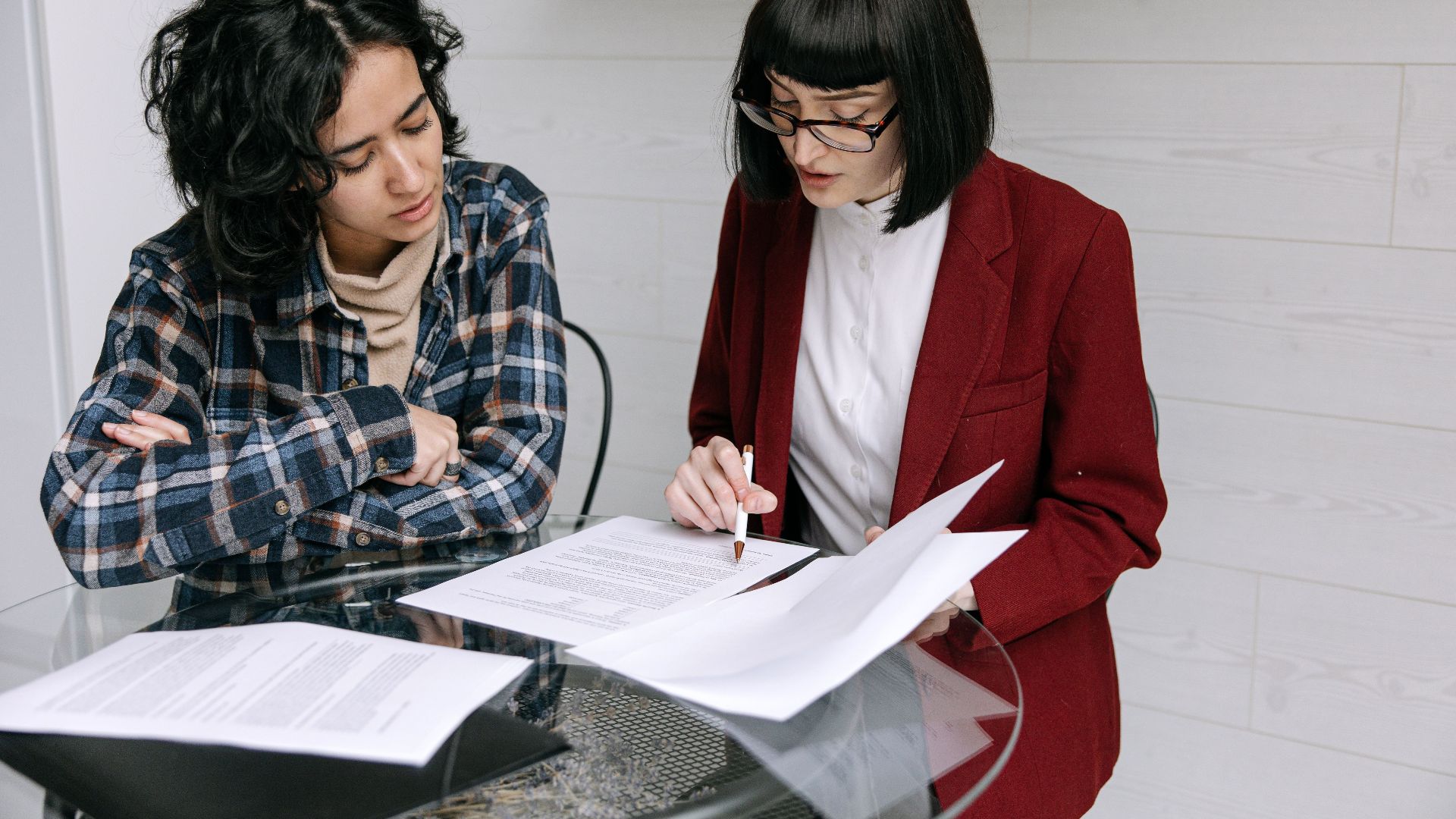 Two women collaborating at a glass table, reviewing business documents in a modern office setting.