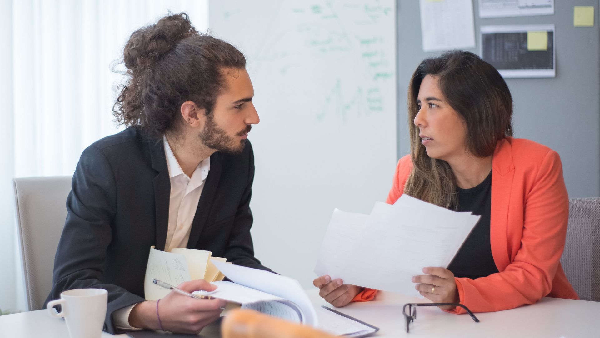 Two colleagues engaged in a discussion at an office table with documents and coffee.