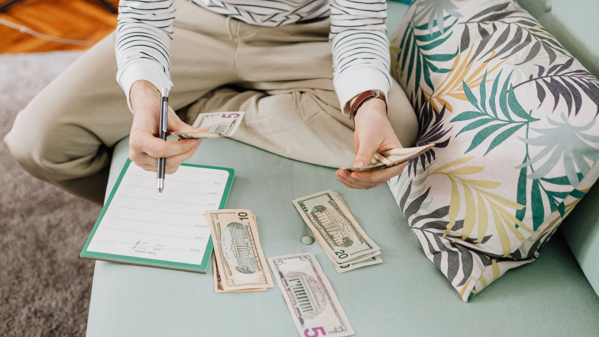A young man wearing a beanie counts cash while sitting on a couch, surrounded by notes and a notebook.