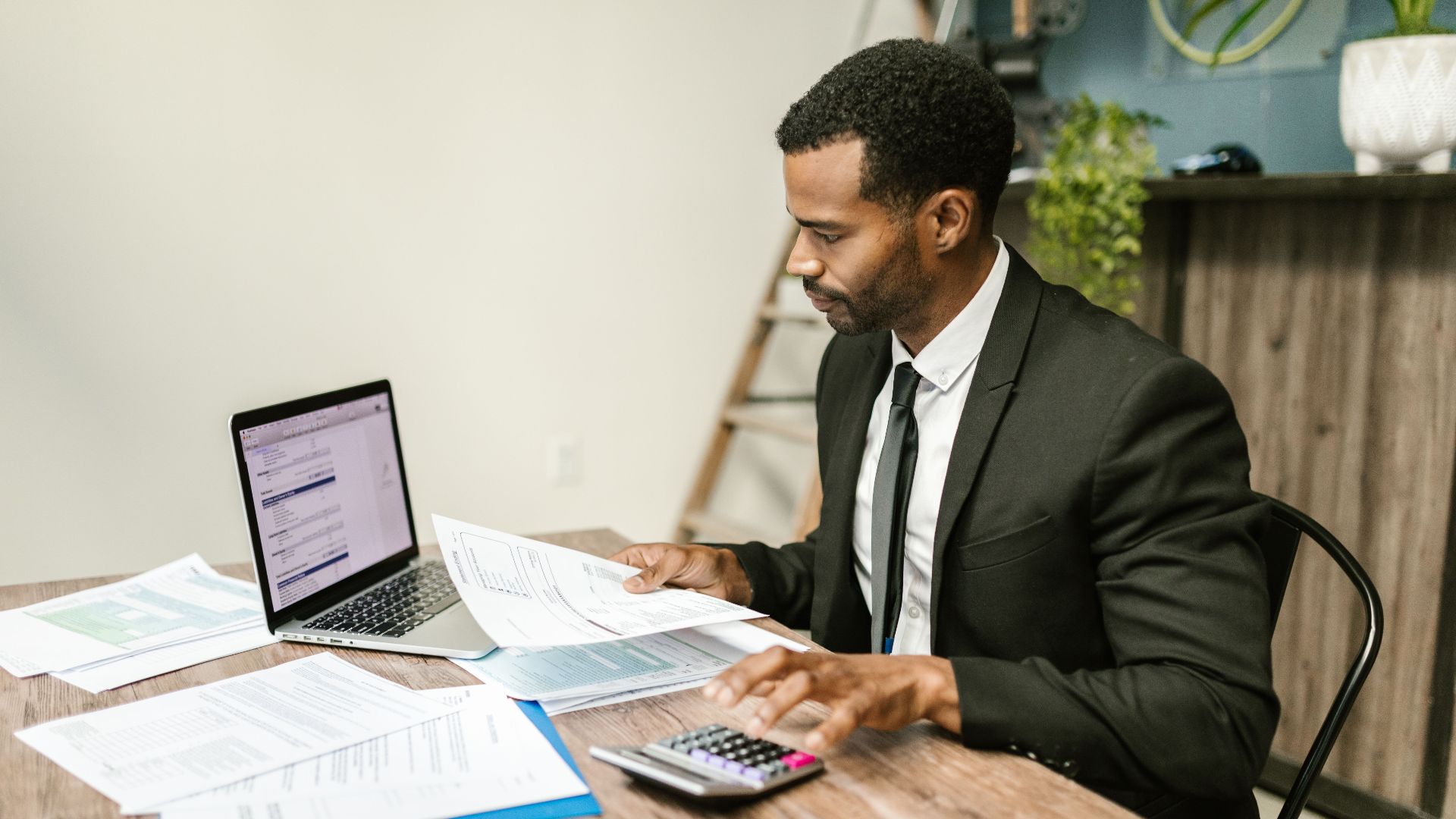 A businessman in a suit working on documents and using a laptop at an office desk.