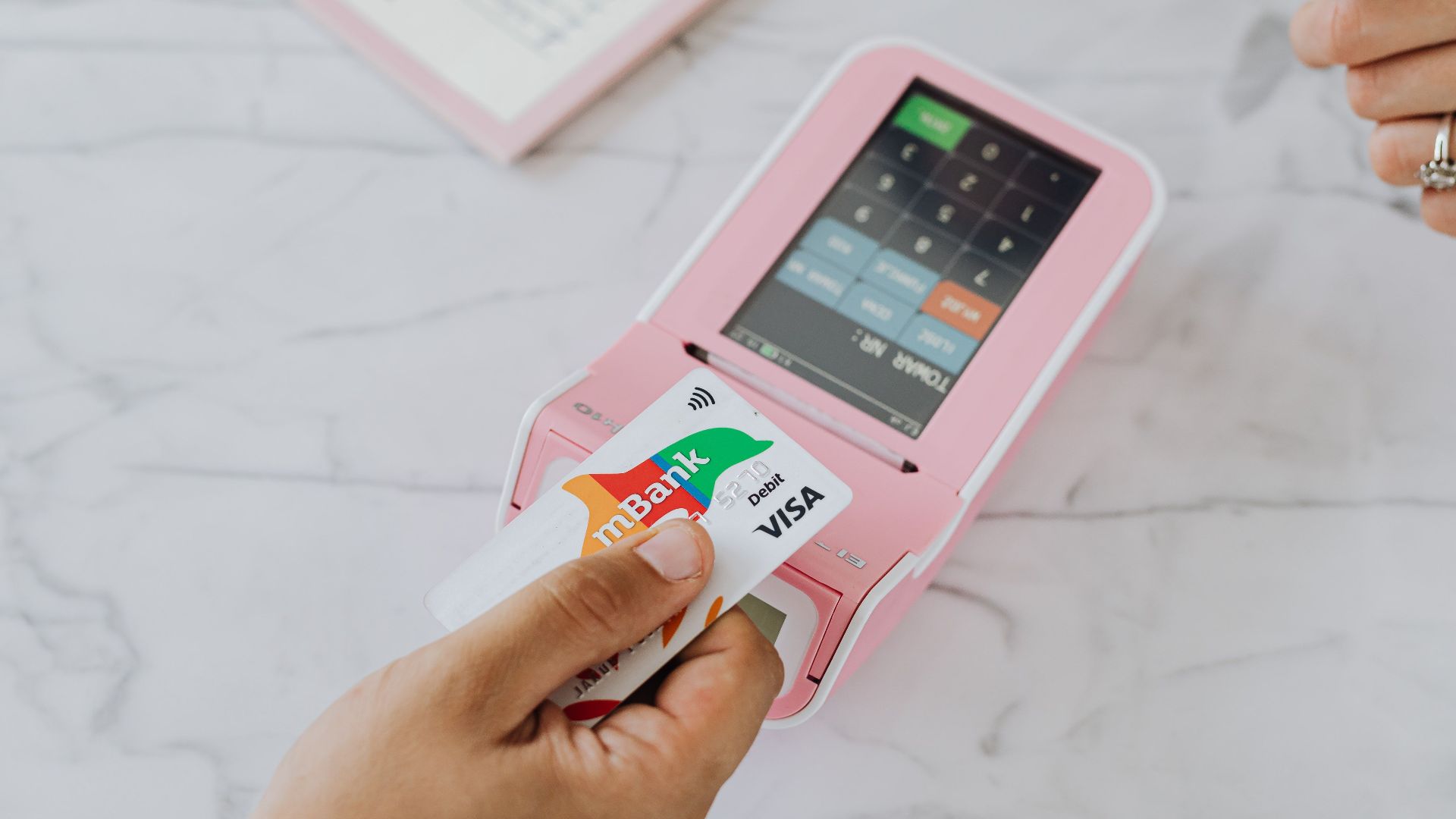 Hand using a contactless card on a pink POS terminal for a wireless transaction.
