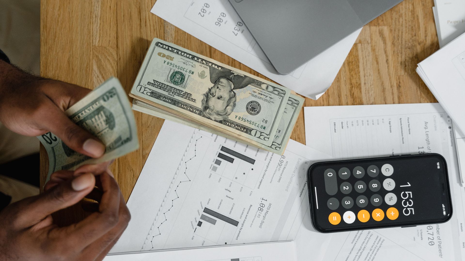 Close-up of hands counting cash on desk with calculator, charts, and laptop, illustrating financial management.