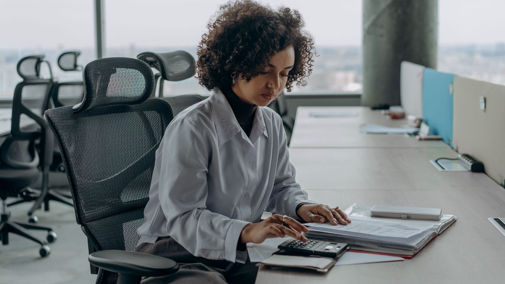 A professional woman with curly hair calculating documents in a modern office setting.