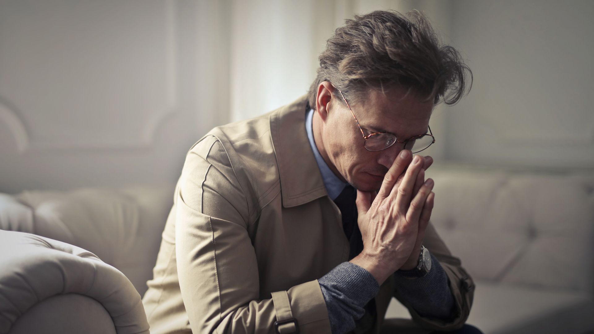 Side view of upset male entrepreneur in formal wear sitting on couch in modern living room and contemplating about financial problems while leaning on hands and looking down
