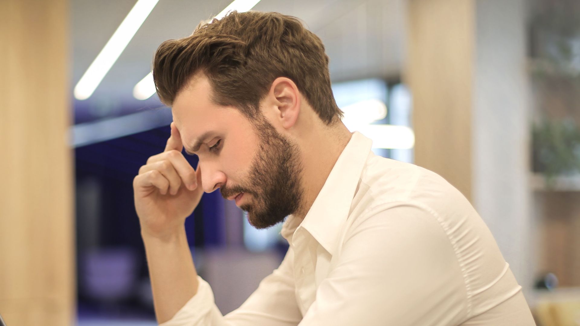 A thoughtful man in a shirt works on his laptop at a modern indoor office space.