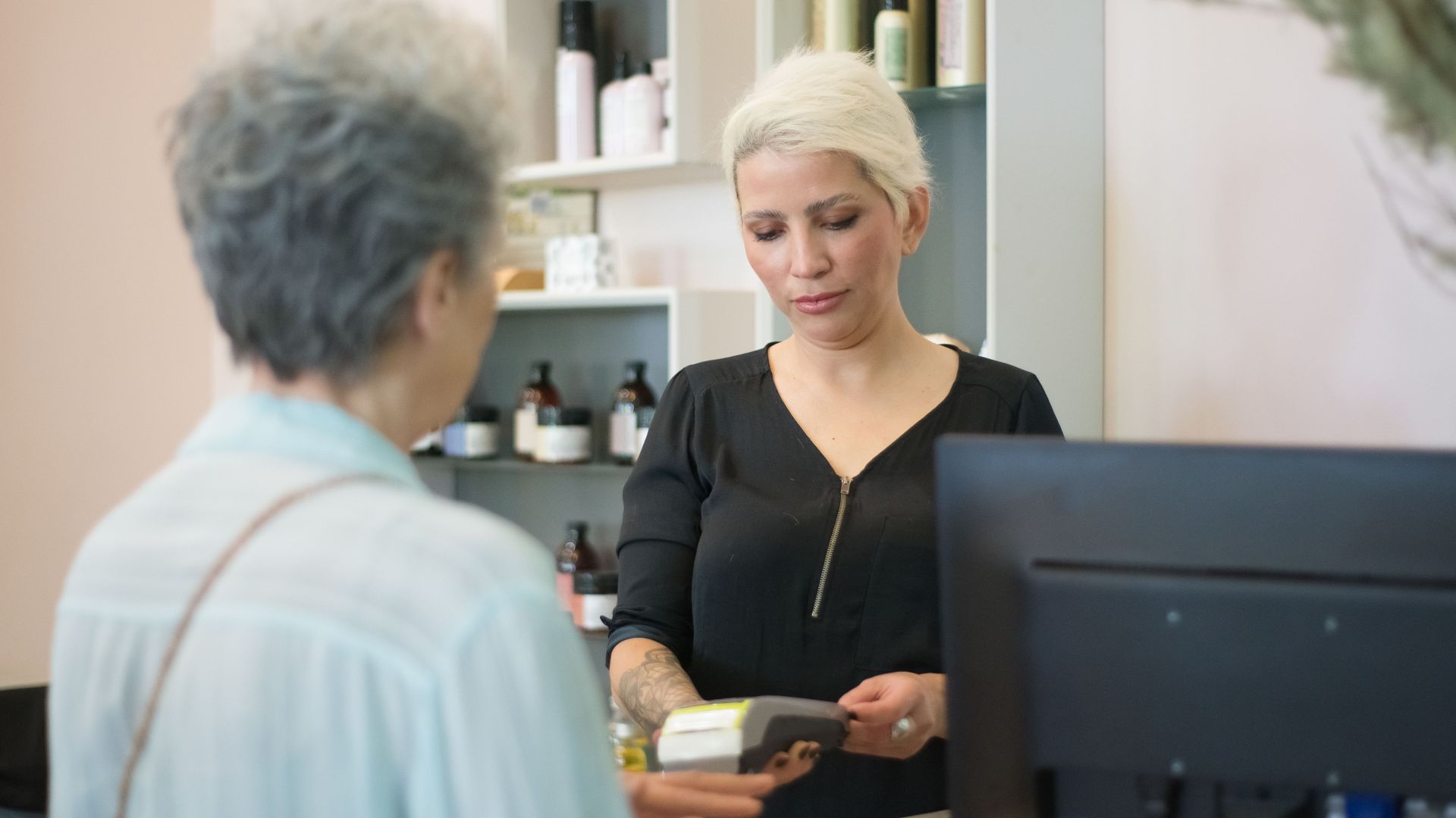 Two women at a hair salon checkout, one making a payment using a card reader.
