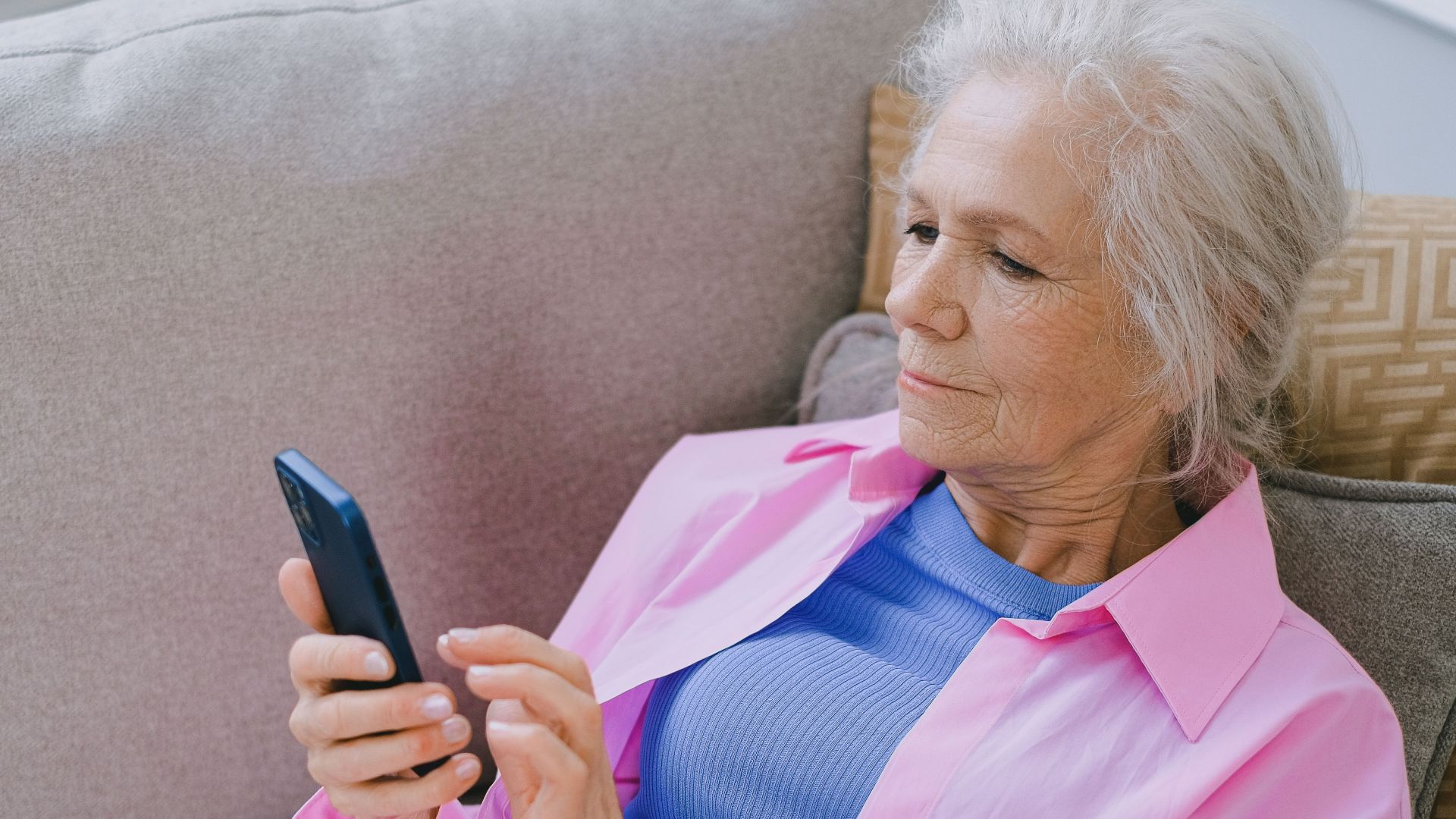 Elderly woman relaxing on sofa while using smartphone for communication.