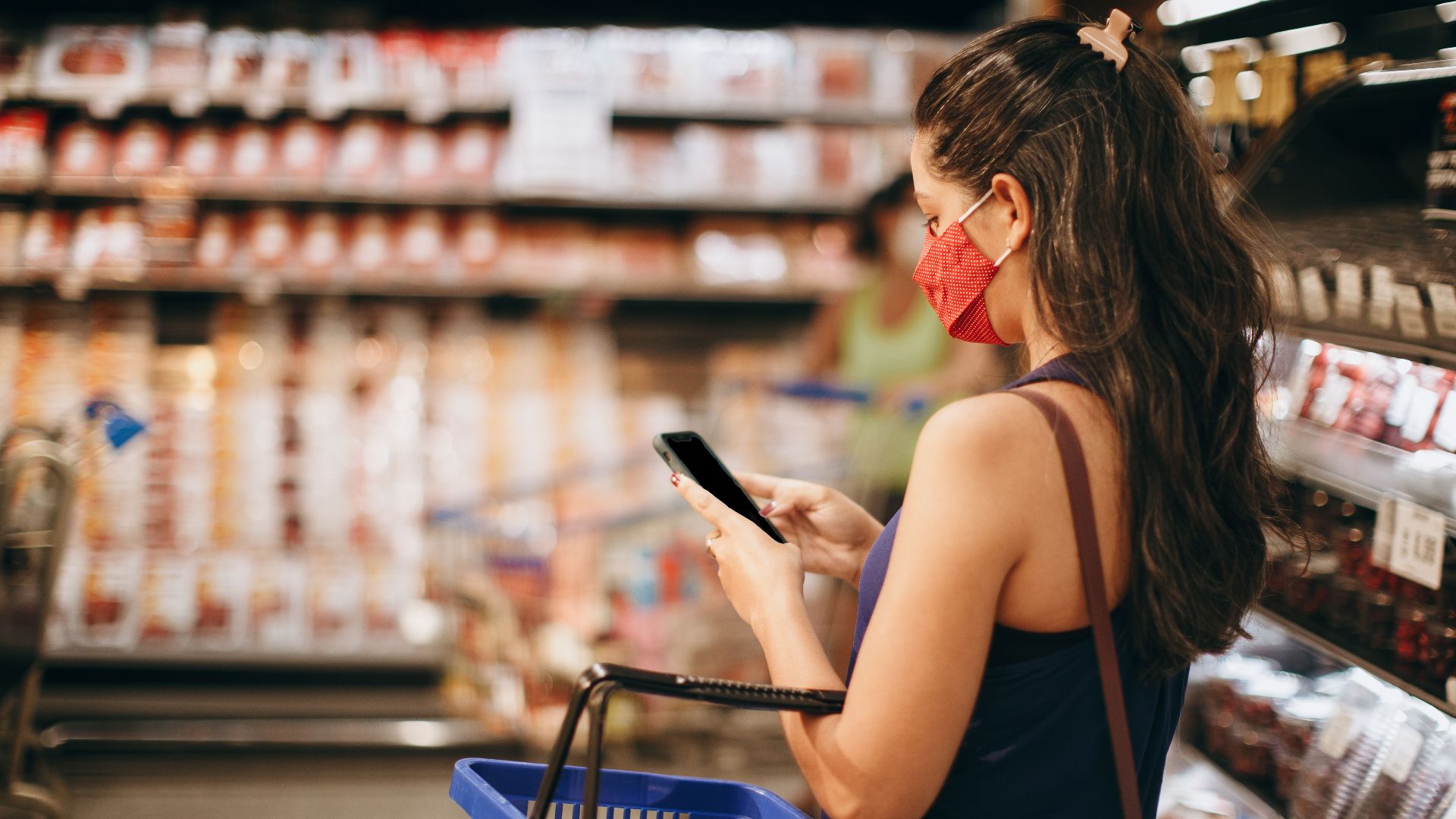 A woman with a mask shops in a grocery store using her smartphone and basket in hand.