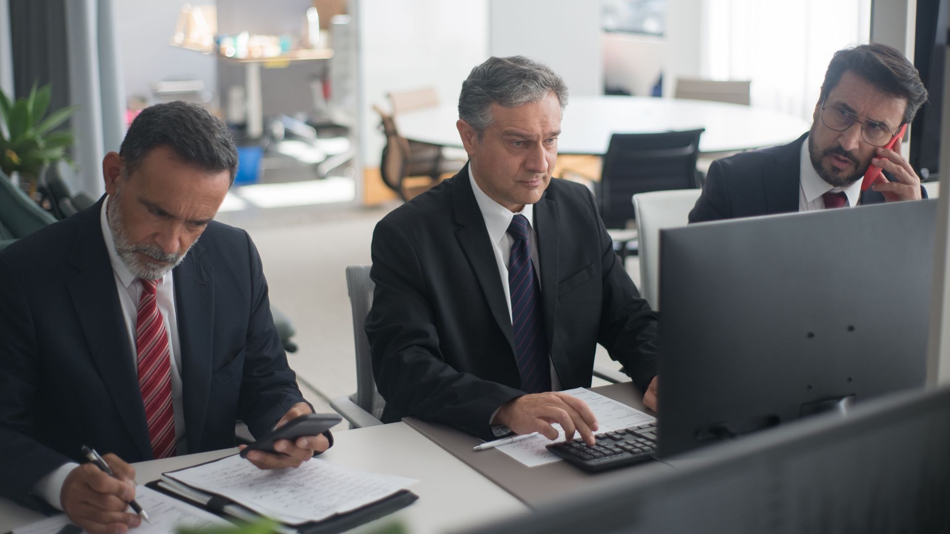 Three businessmen in suits working on computers and documents in a modern office setting.