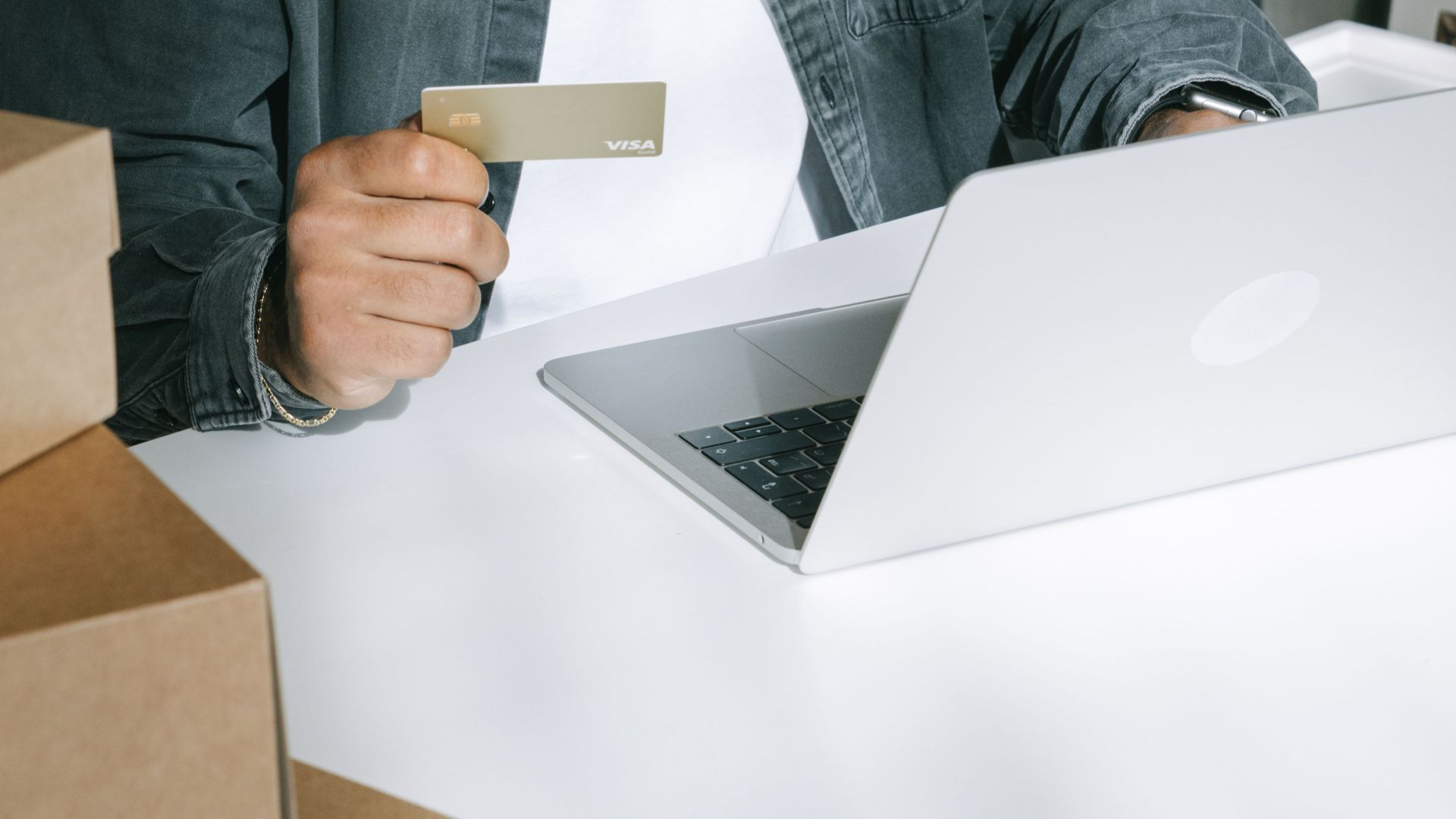 Person using a credit card for online shopping on a laptop surrounded by shipping boxes.