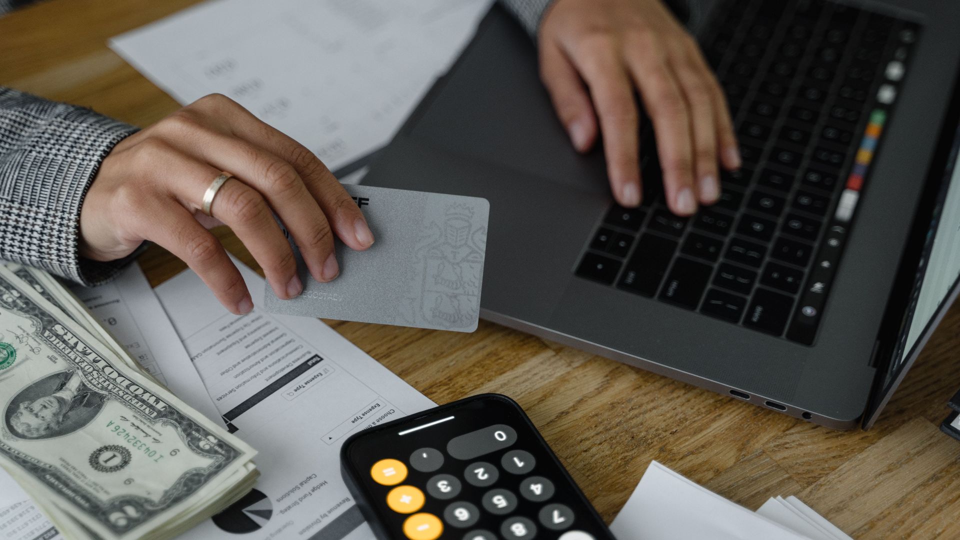 Close-up of a person using a credit card and laptop in a financial setting with cash and calculator.