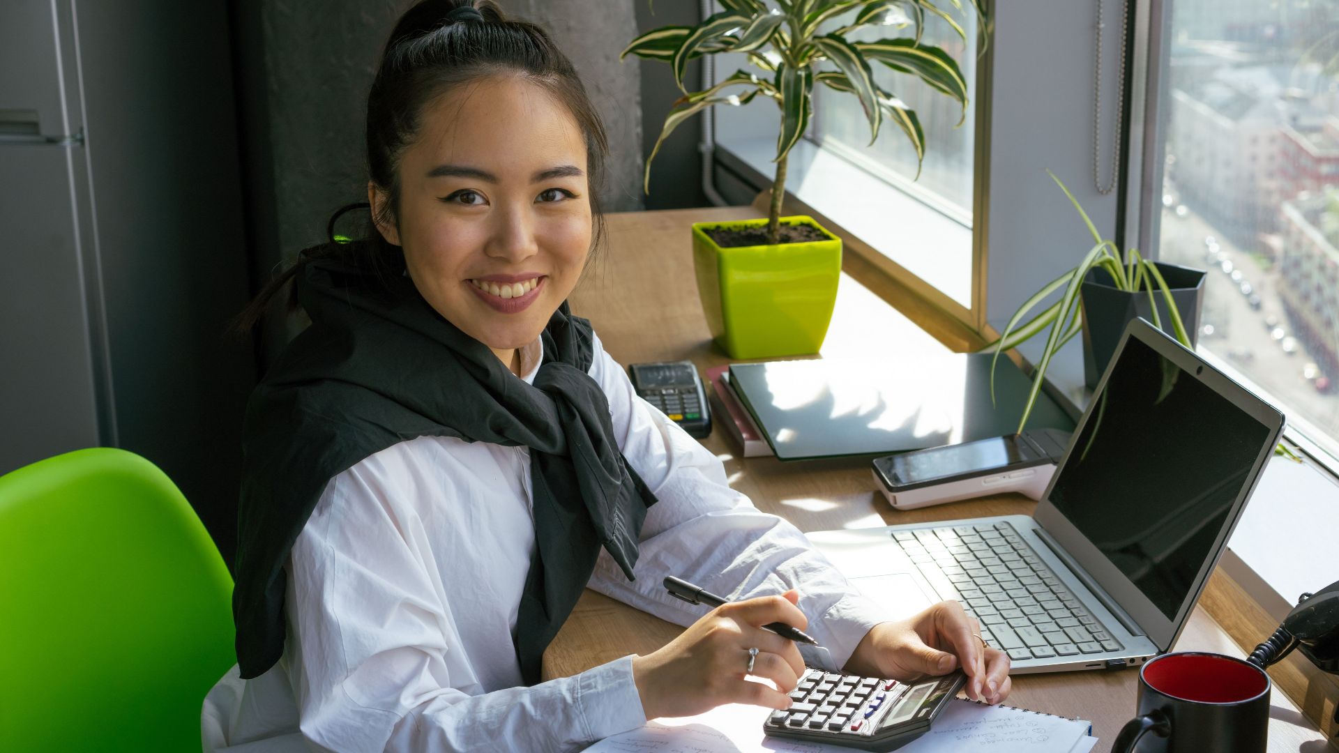 Confident woman working with calculator in bright office setting, surrounded by office tools.