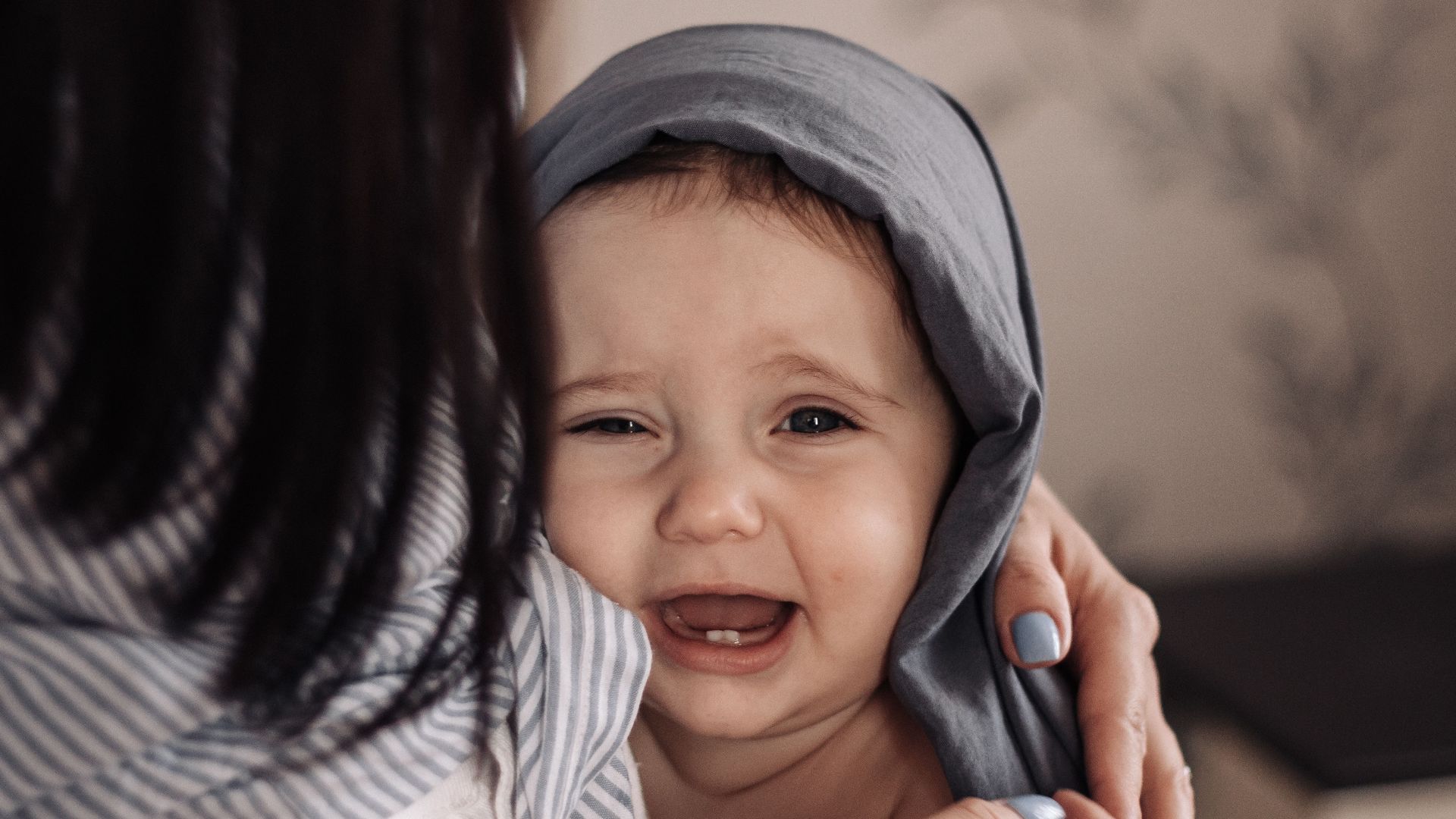Emotional baby wrapped in a grey hood, captured indoors, exhibiting a charming crying face.