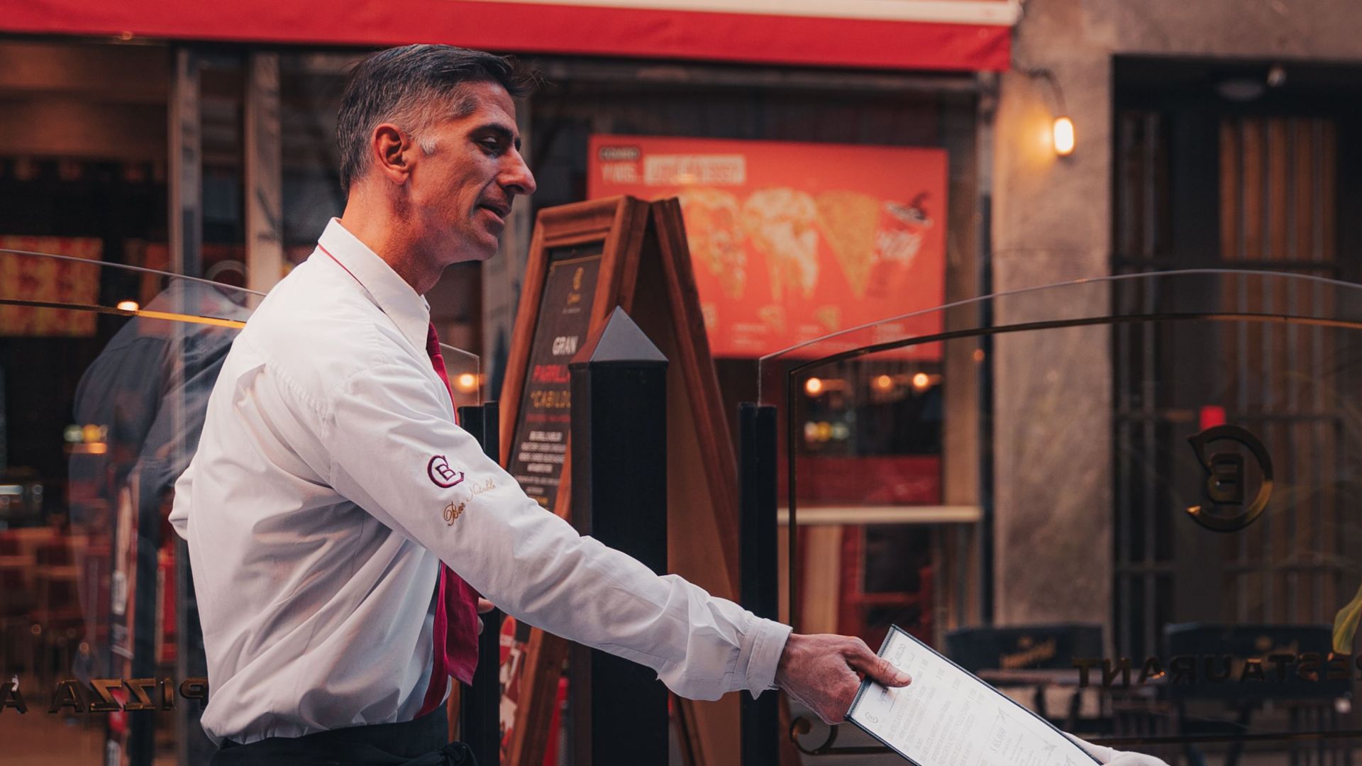 A waiter serves a menu to a seated customer at an outdoor restaurant with colorful signage.
