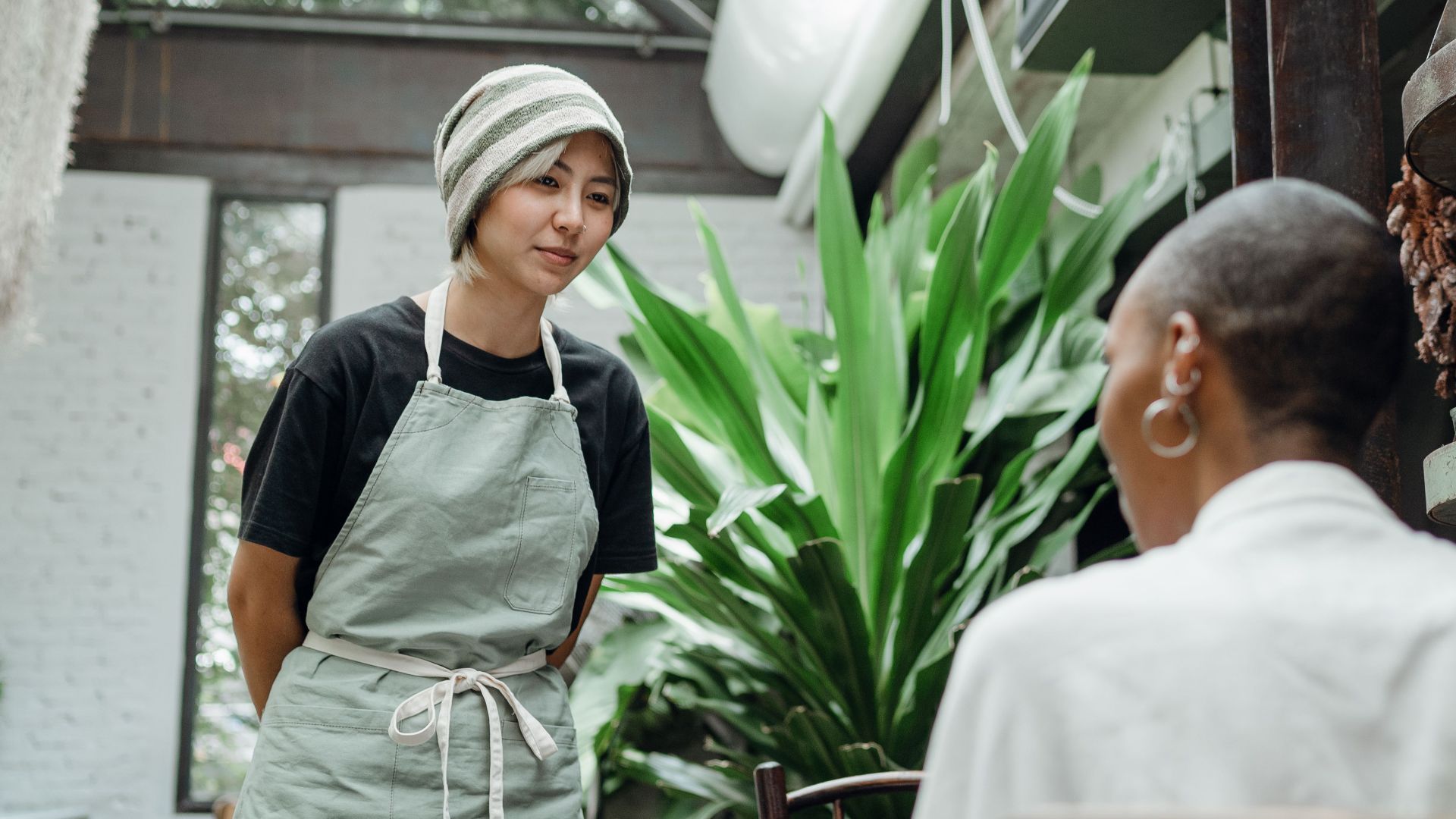 Friendly young Asian waitress in apron and hat standing near table and talking to female customer while working in cozy cafe