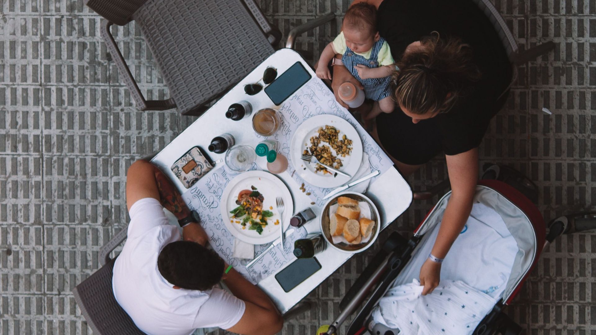 A family enjoys a meal at an outdoor restaurant table, viewed from above with a baby in a stroller.
