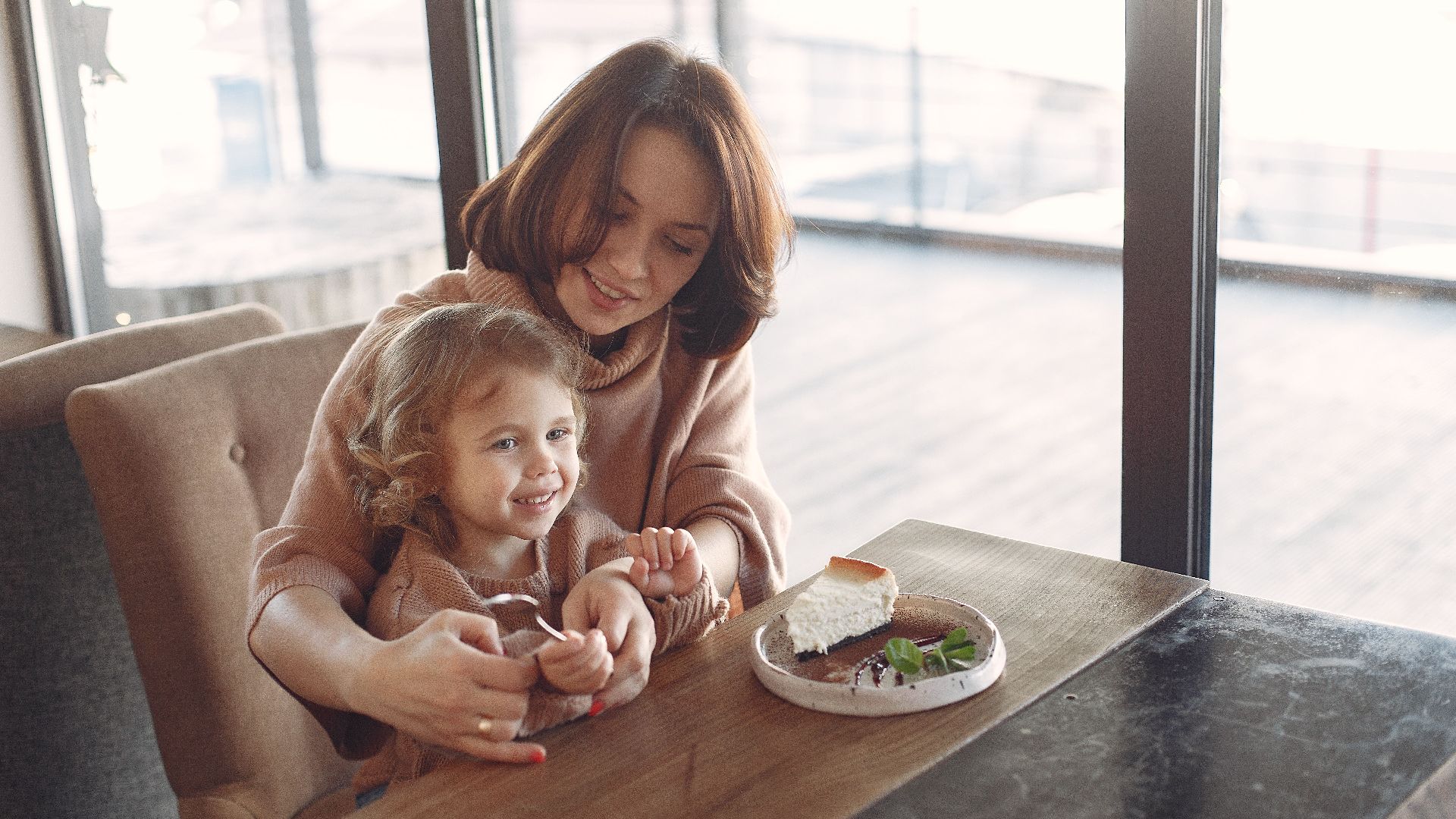 Young woman in casual clothes sitting at table and eating piece of cake with little girl while spending time at cafeteria together