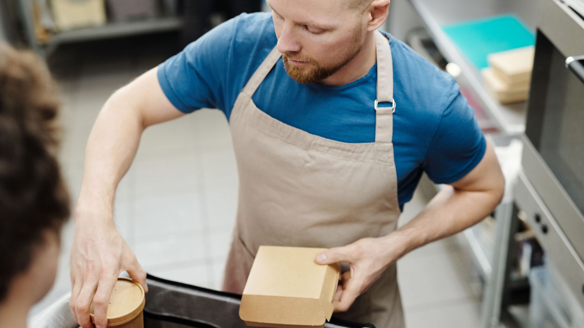 A man wearing an apron packs food boxes in a kitchen setting.