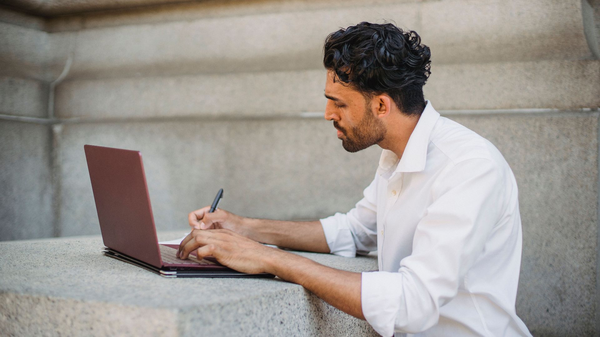 A man writing on a laptop outdoors, seated on a stone surface and focused on work.