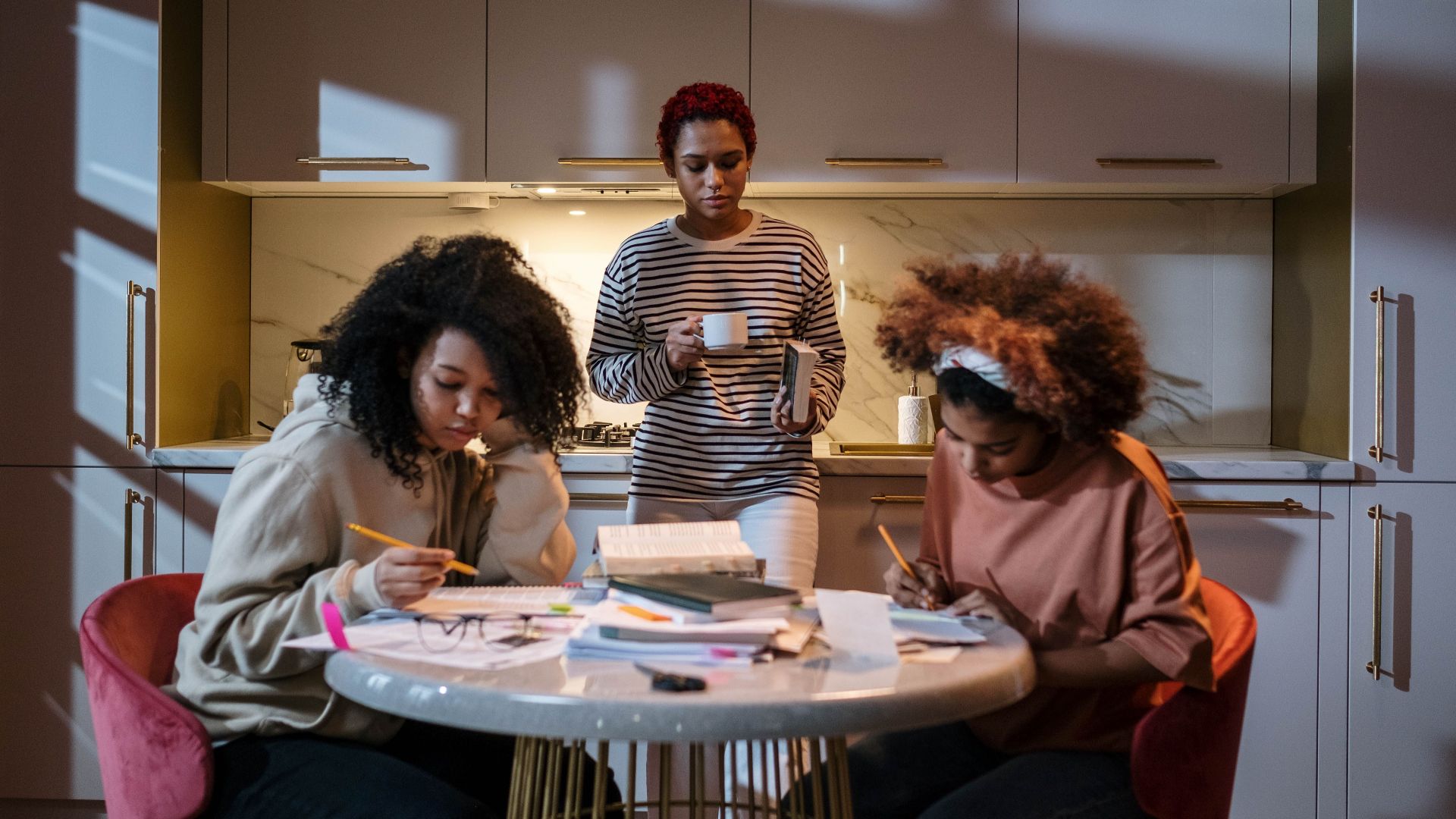 Three young women studying and enjoying coffee in a cozy kitchen, focused and engaged.