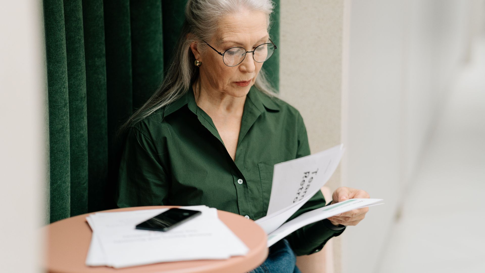 Elderly woman with gray hair reading papers at a small table indoors, focused and thoughtful.