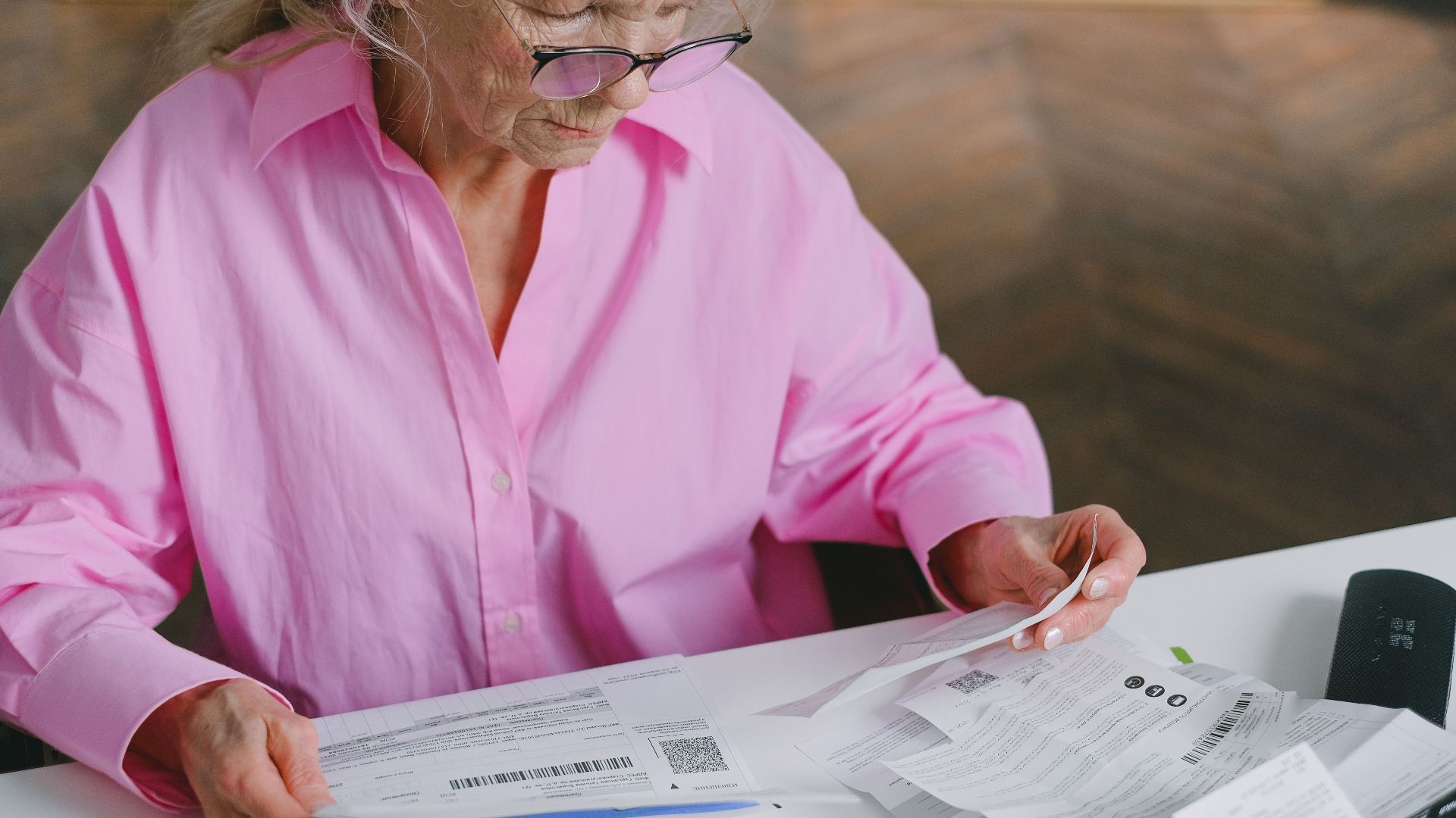 Elderly woman in pink blouse reading documents at a table indoors.