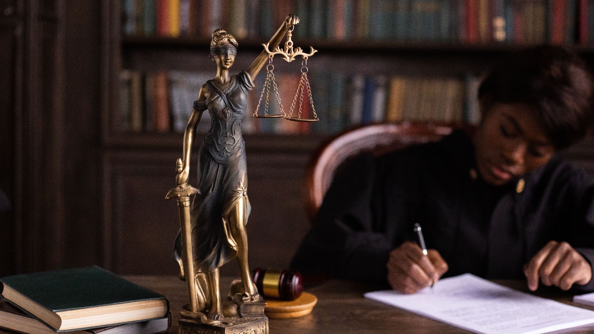 A focused judge writing on documents beside a Lady Justice statue in an office.