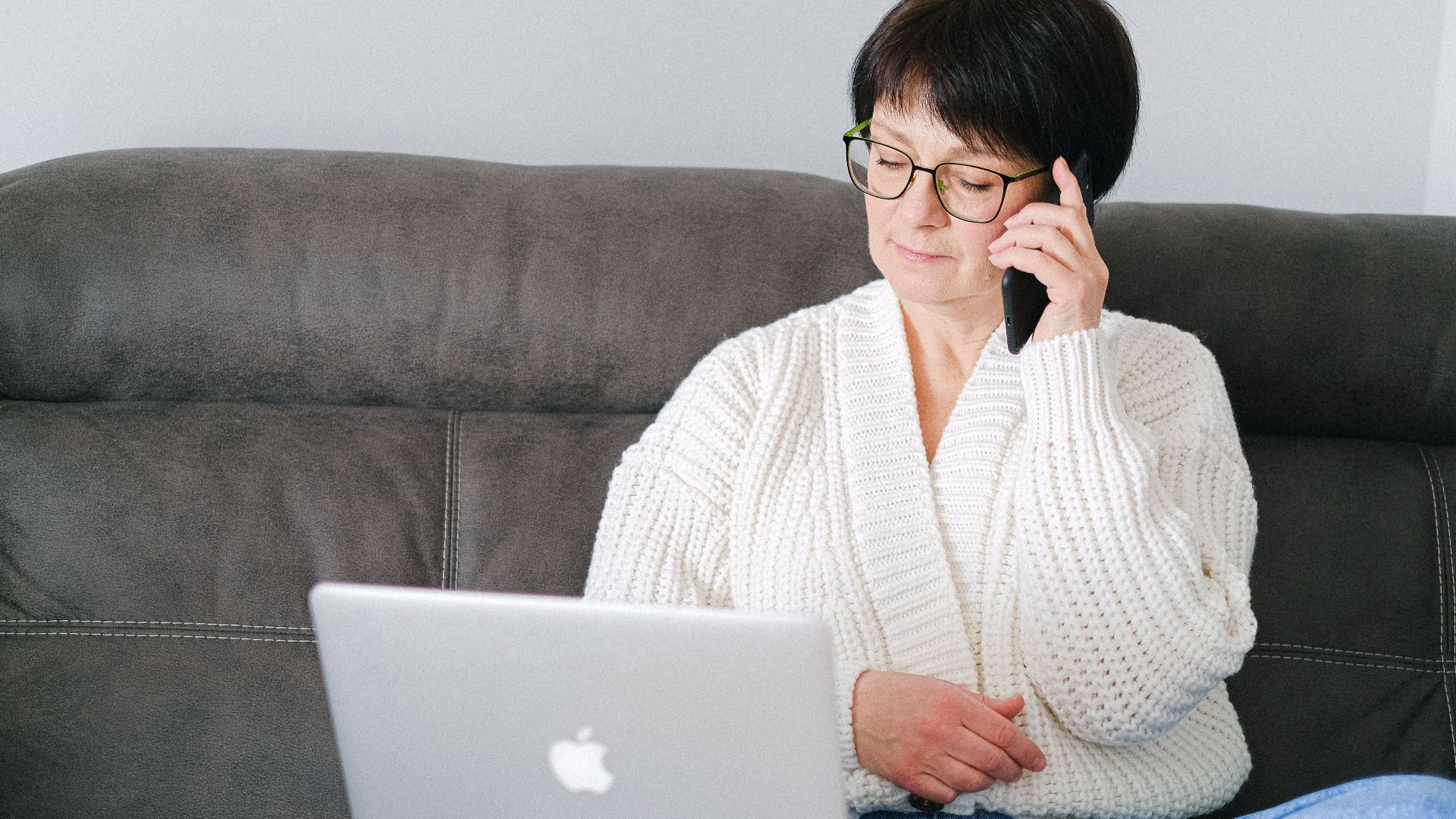 Elderly woman sitting on a sofa, using a laptop and phone simultaneously.