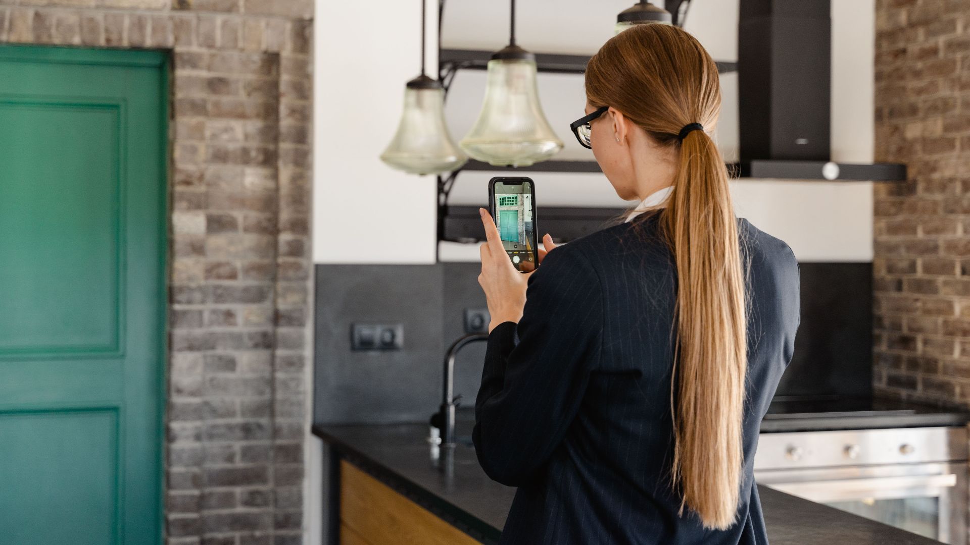 Rear view of a woman with long hair taking a photo with a smartphone in a stylish kitchen.