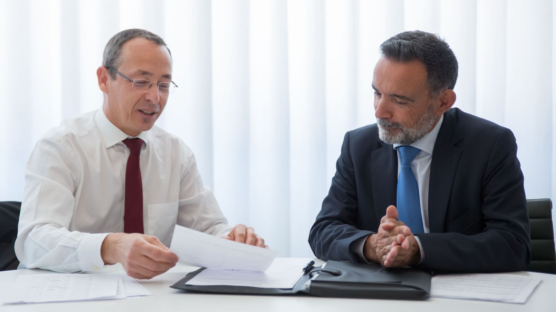 Two businessmen in suits having a discussion over documents in a bright office setting.