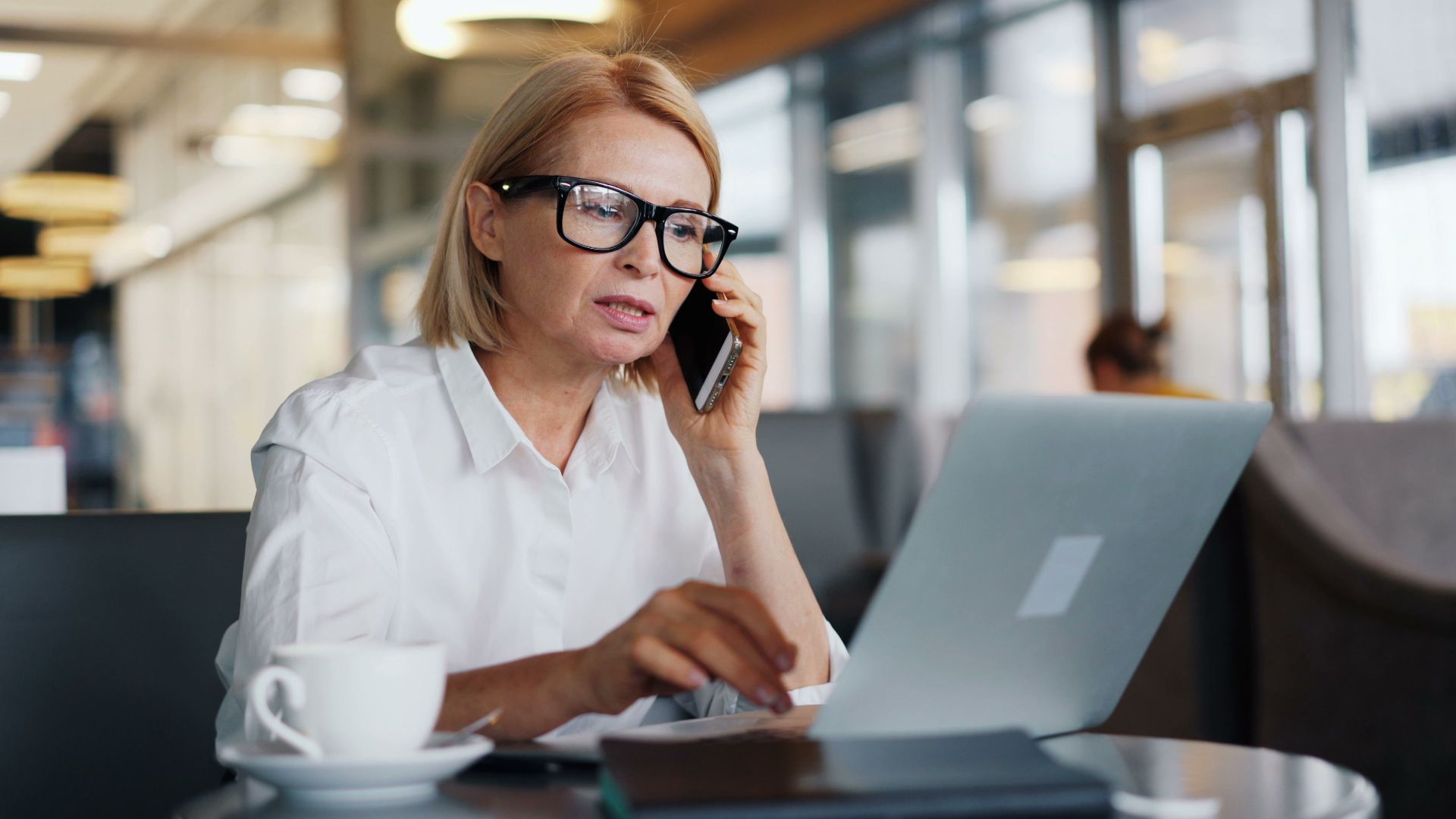 Mature woman in a café using a laptop and smartphone for business communication.