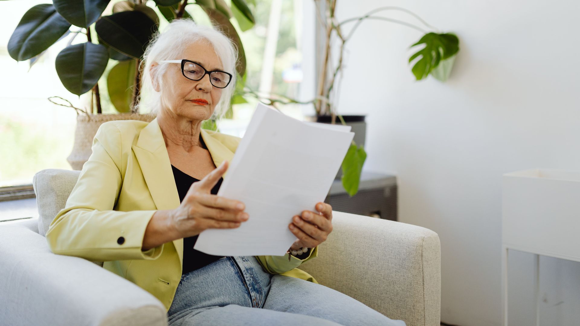 Elderly woman in yellow blazer reading papers at home near plants.
