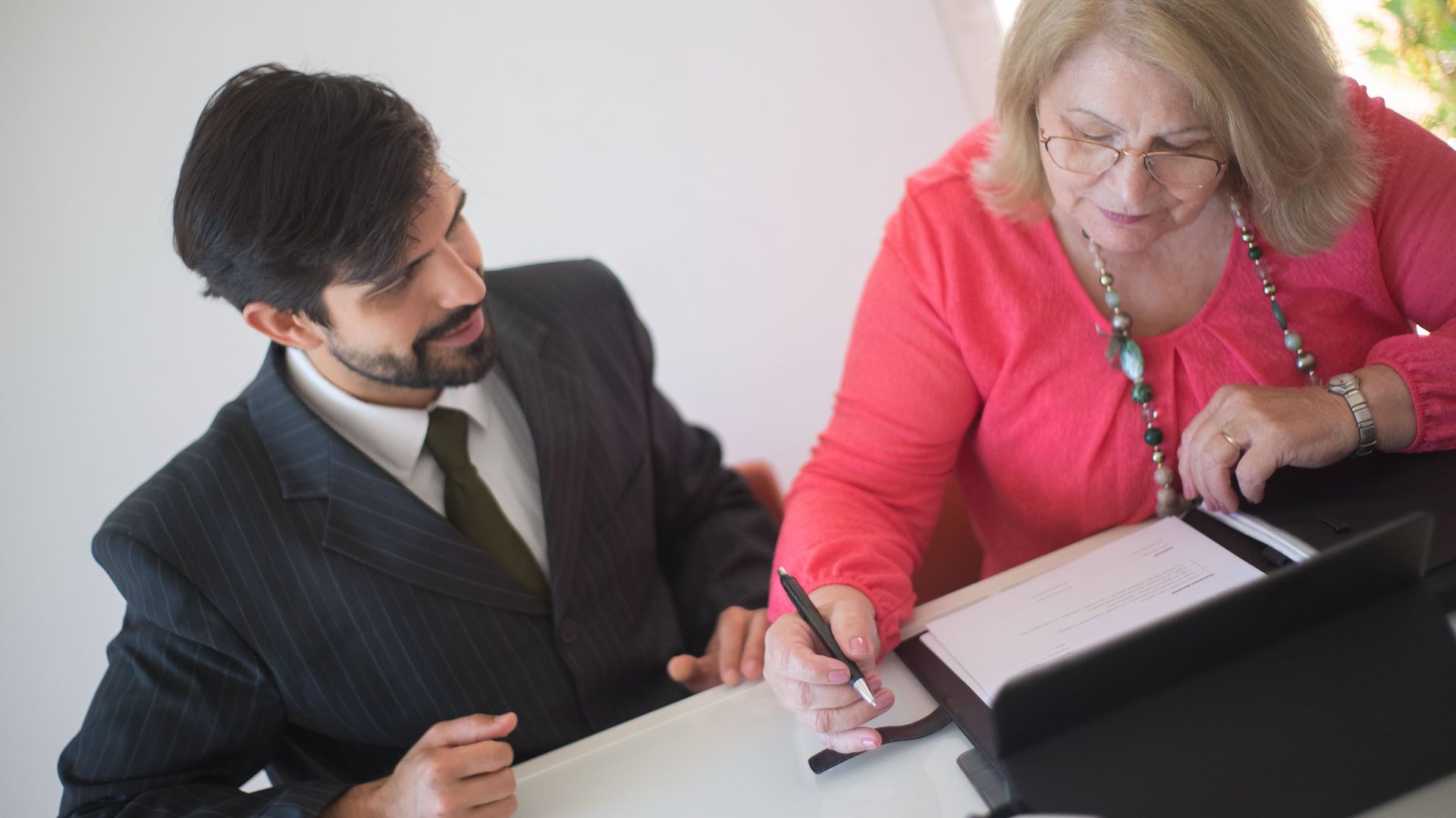 A senior woman signing paperwork with a young professional assisting her at a desk.