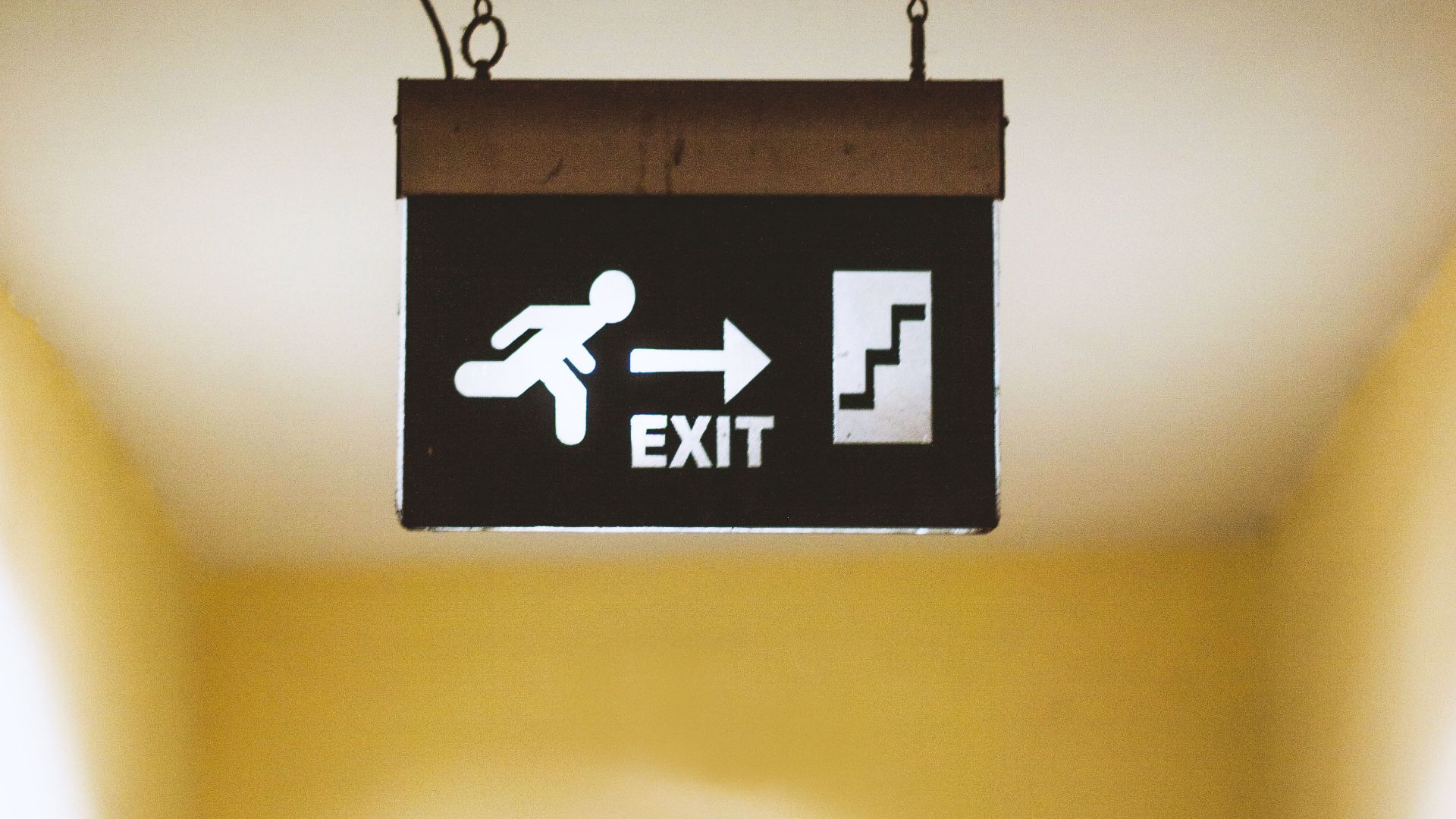A bright exit sign hanging indoors with arrow and man running symbol pointing to stairs.