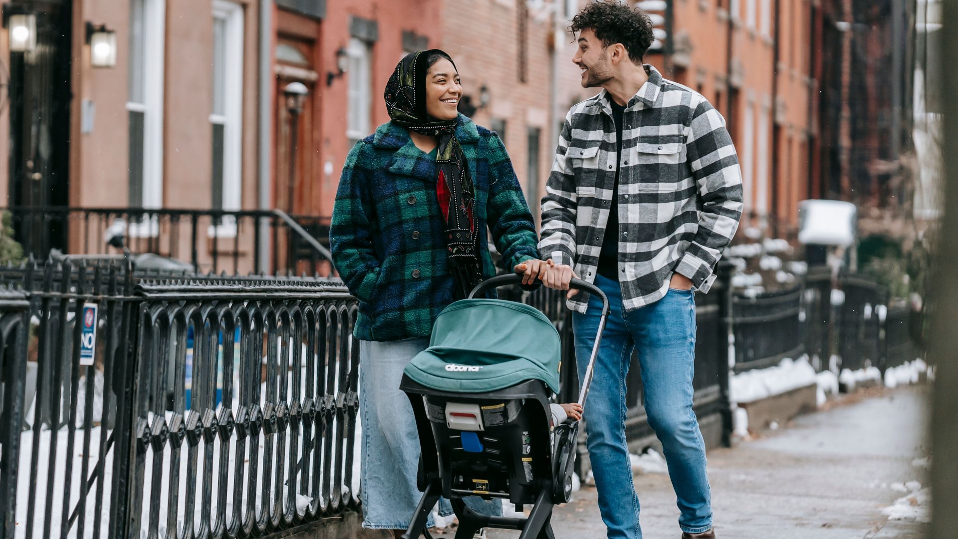 Full body of multiracial parents looking at each other while strolling on sidewalk along buildings with anonymous child in baby carriage