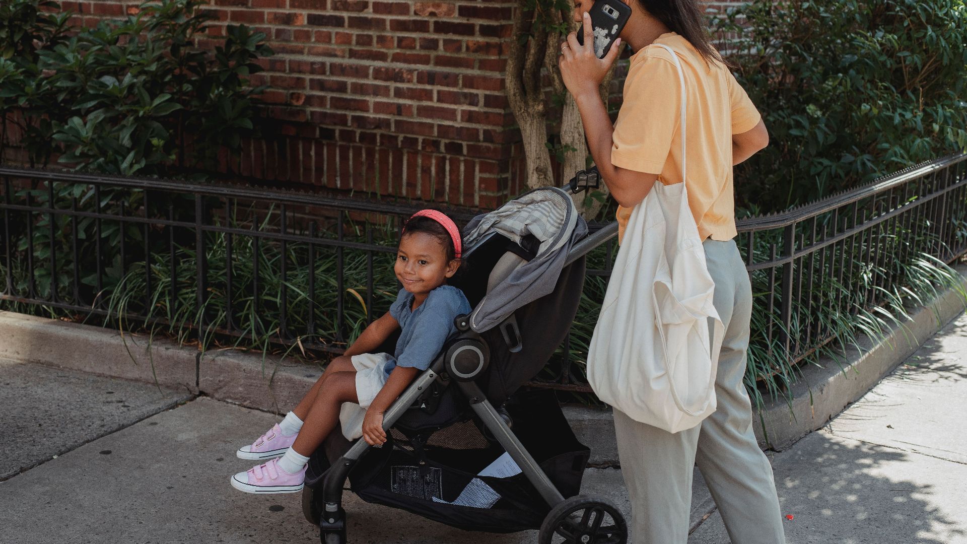 Crop Asian woman talking on smartphone while pushing baby stroller with little girl during walk on pavement in sunny city