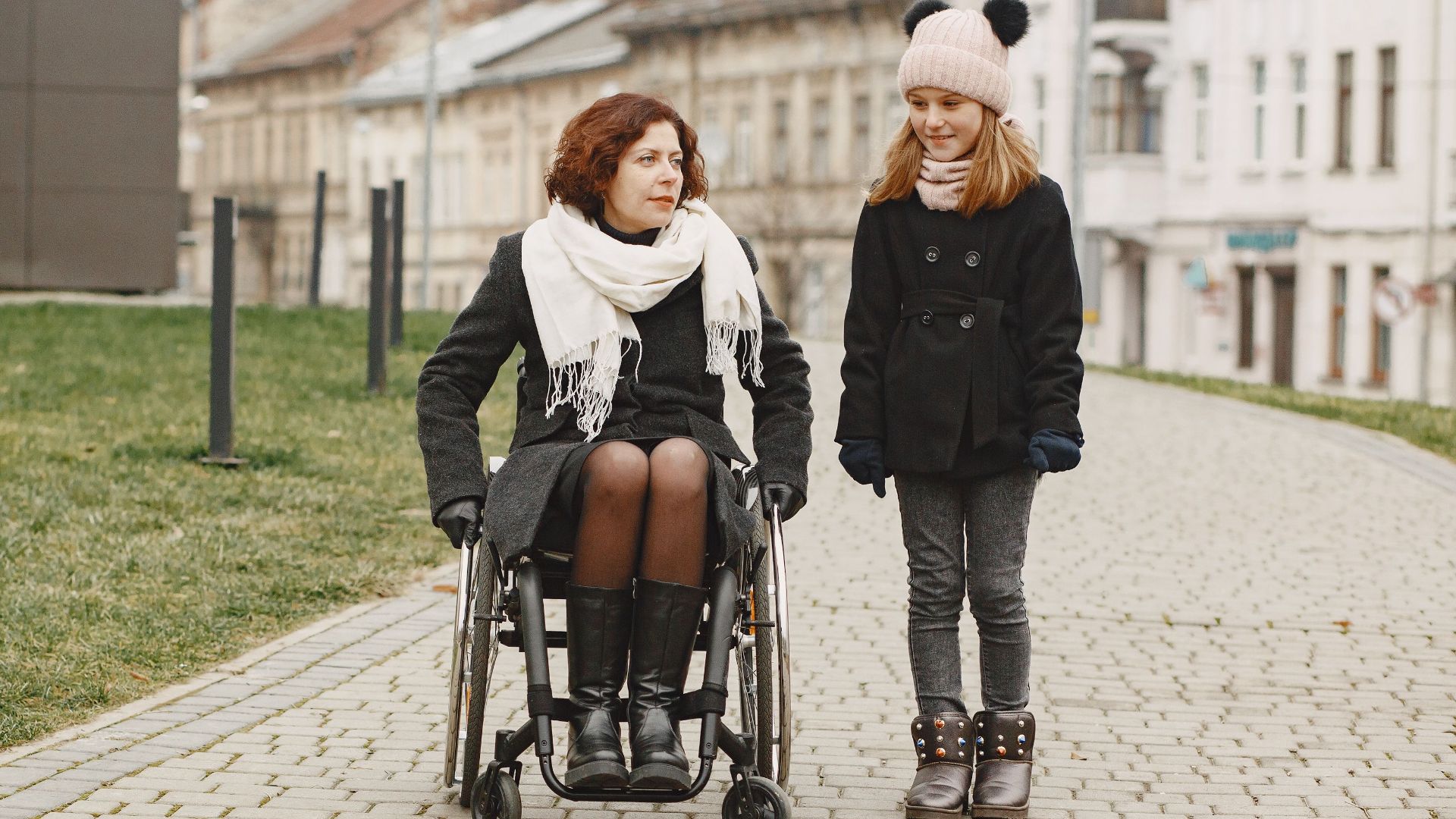 A woman in a wheelchair and a girl walking on a cobbled path in urban setting.