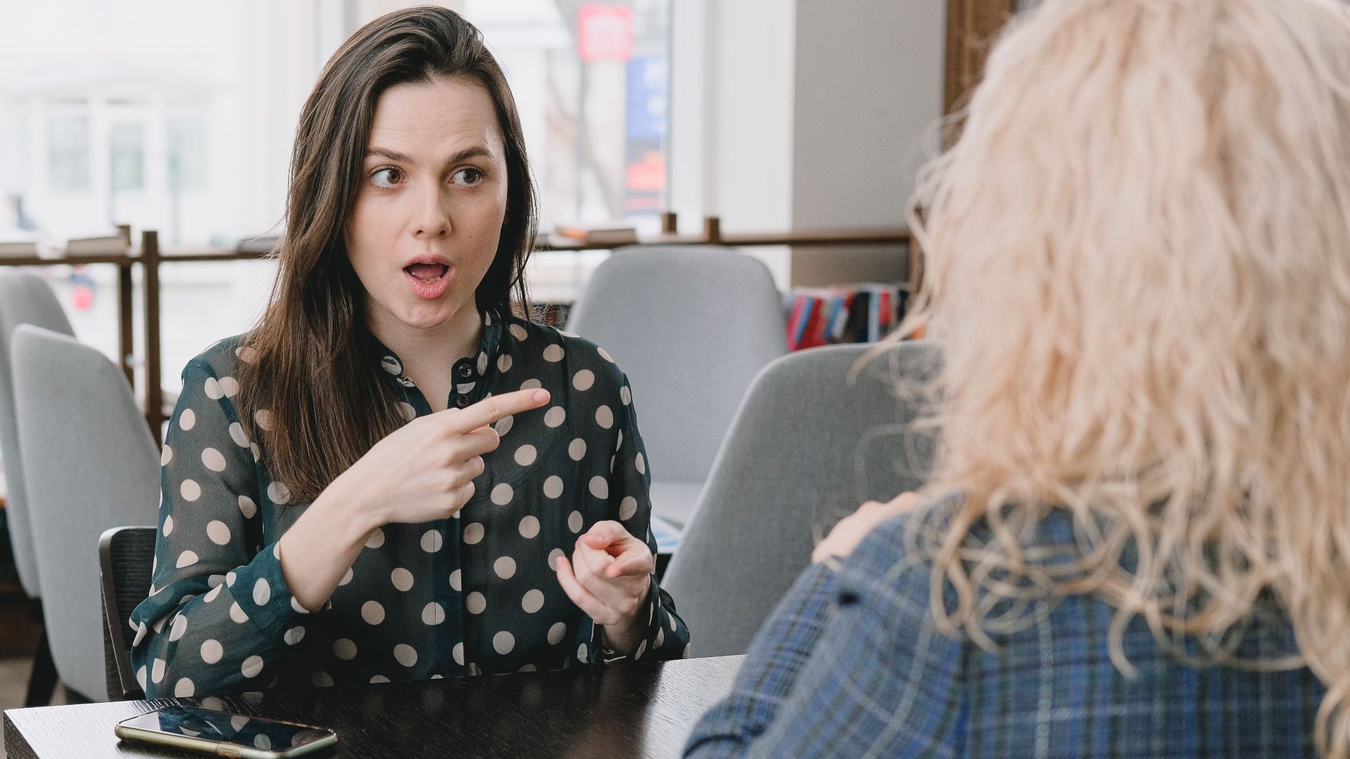 Astonished female pointing away while sitting at table with smartphone and tablet and talking to woman
