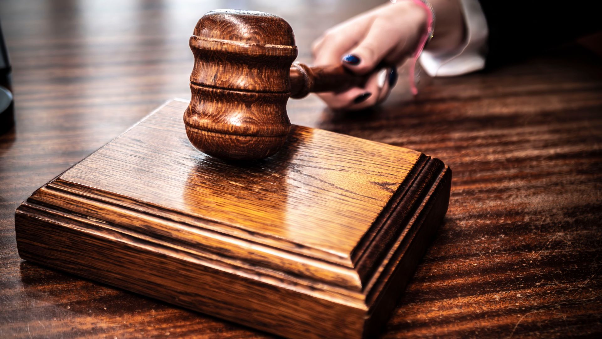 Close-up image of a wooden gavel held by hand on a desk in a courtroom setting.