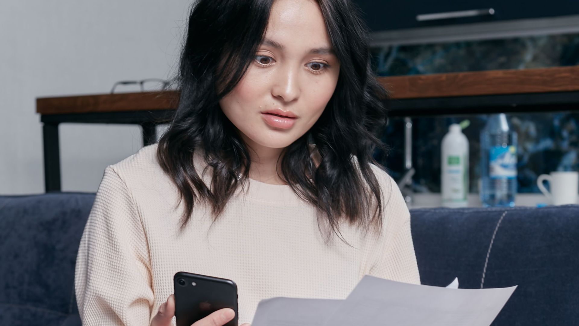 Young woman reviews documents and phone indoors, showcasing concern while seated on a couch with scattered papers. Business concept.