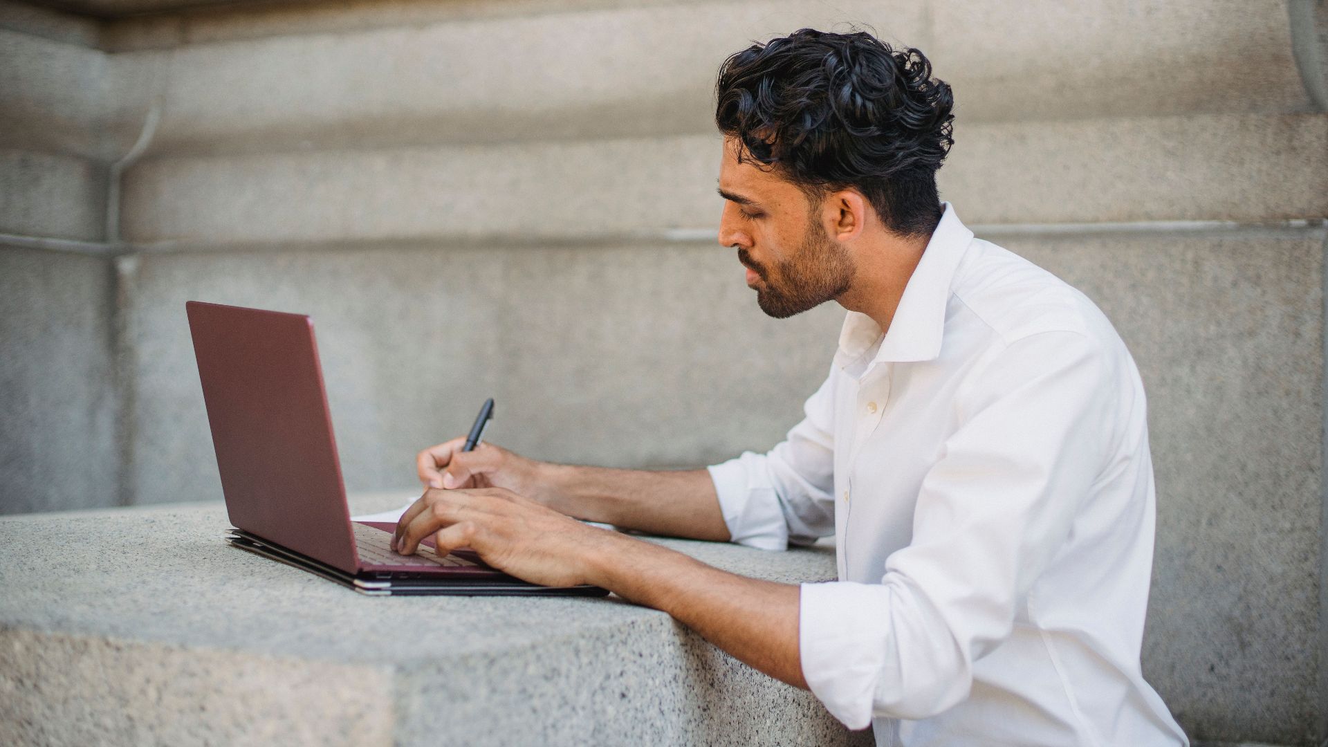 A man writing on a laptop outdoors, seated on a stone surface and focused on work.