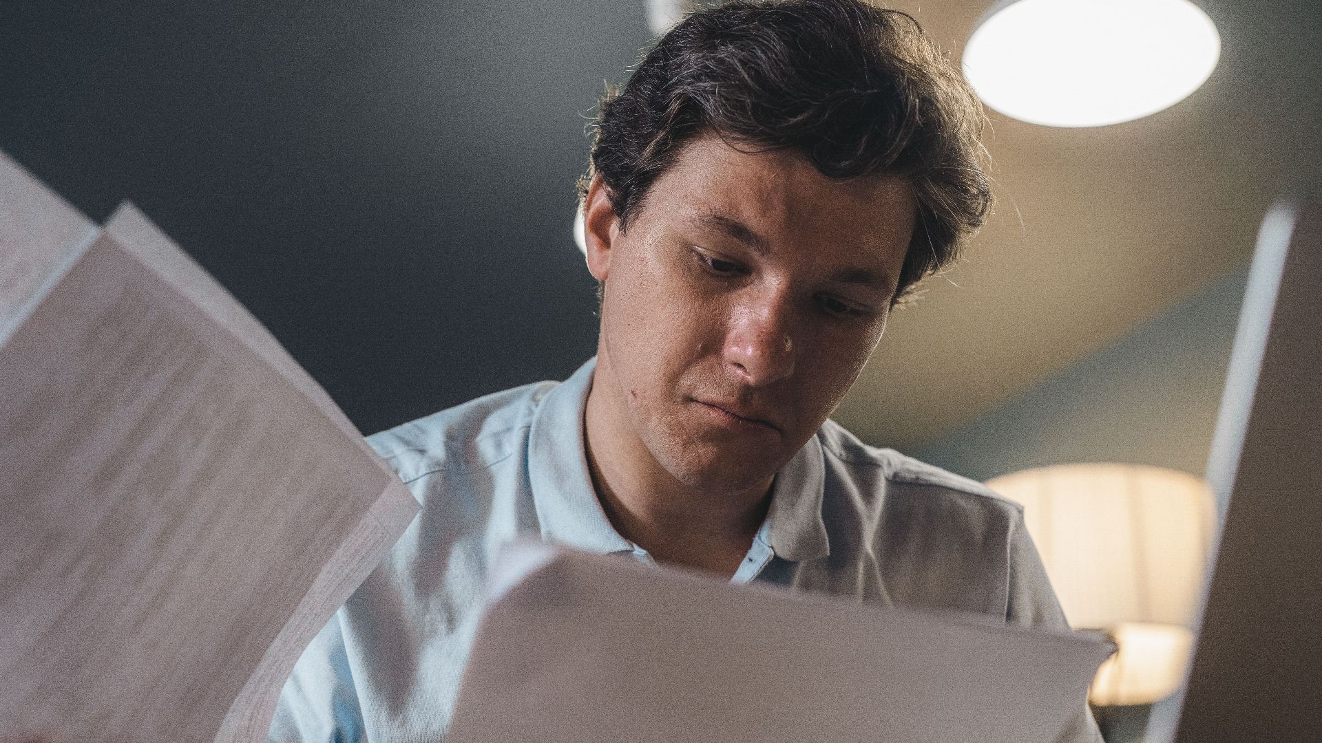 Serious man wearing a light blue shirt intently reading printed papers indoors under warm lighting.