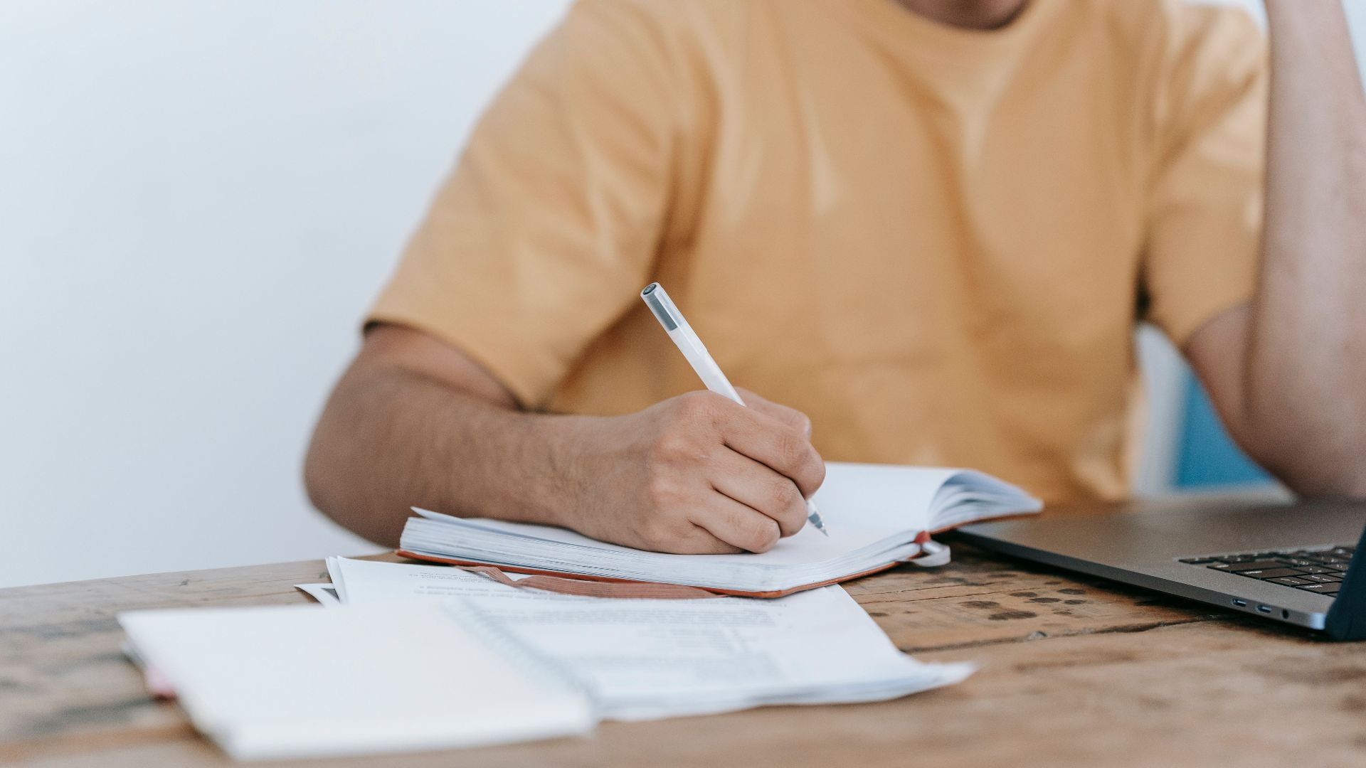 Crop anonymous male in casual wear working on netbook and taking notes in notepad while sitting at desk