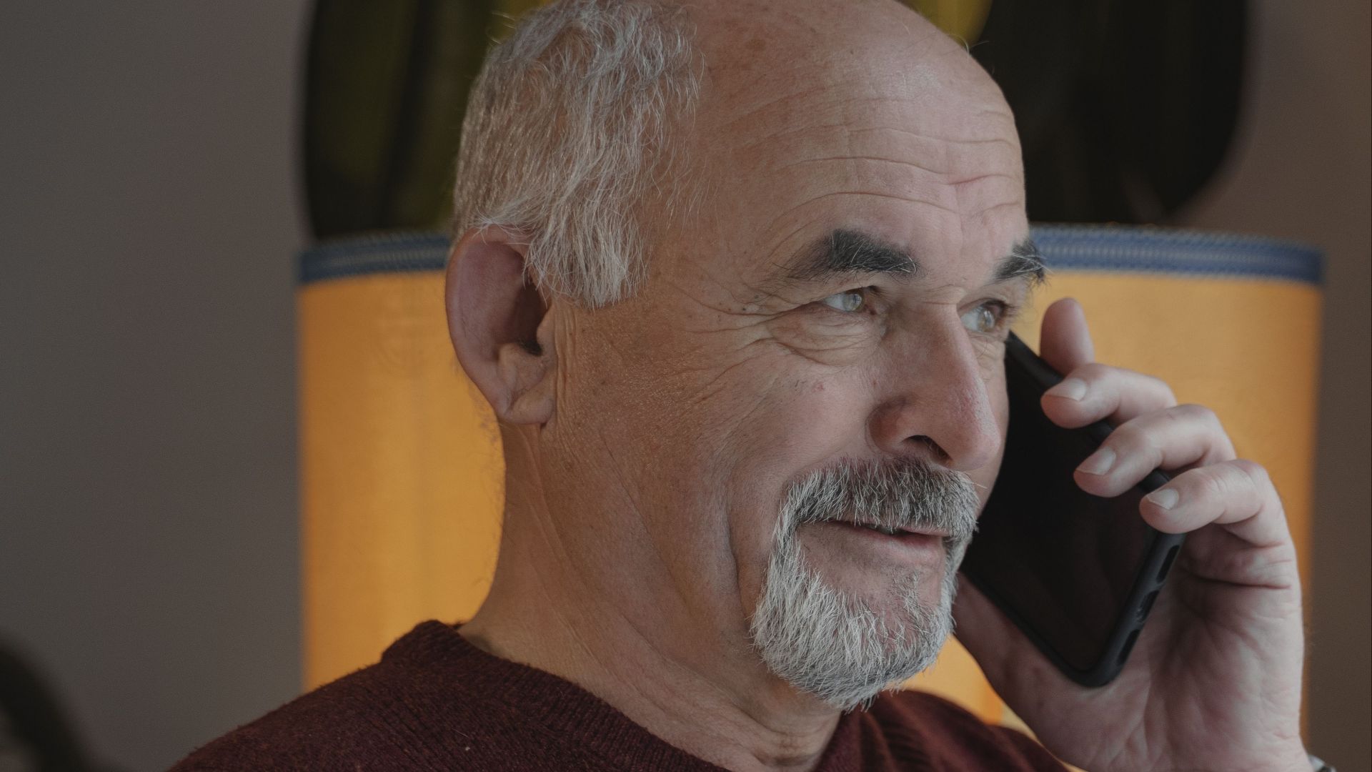 Elderly man making a phone call indoors, wearing a red sweater.
